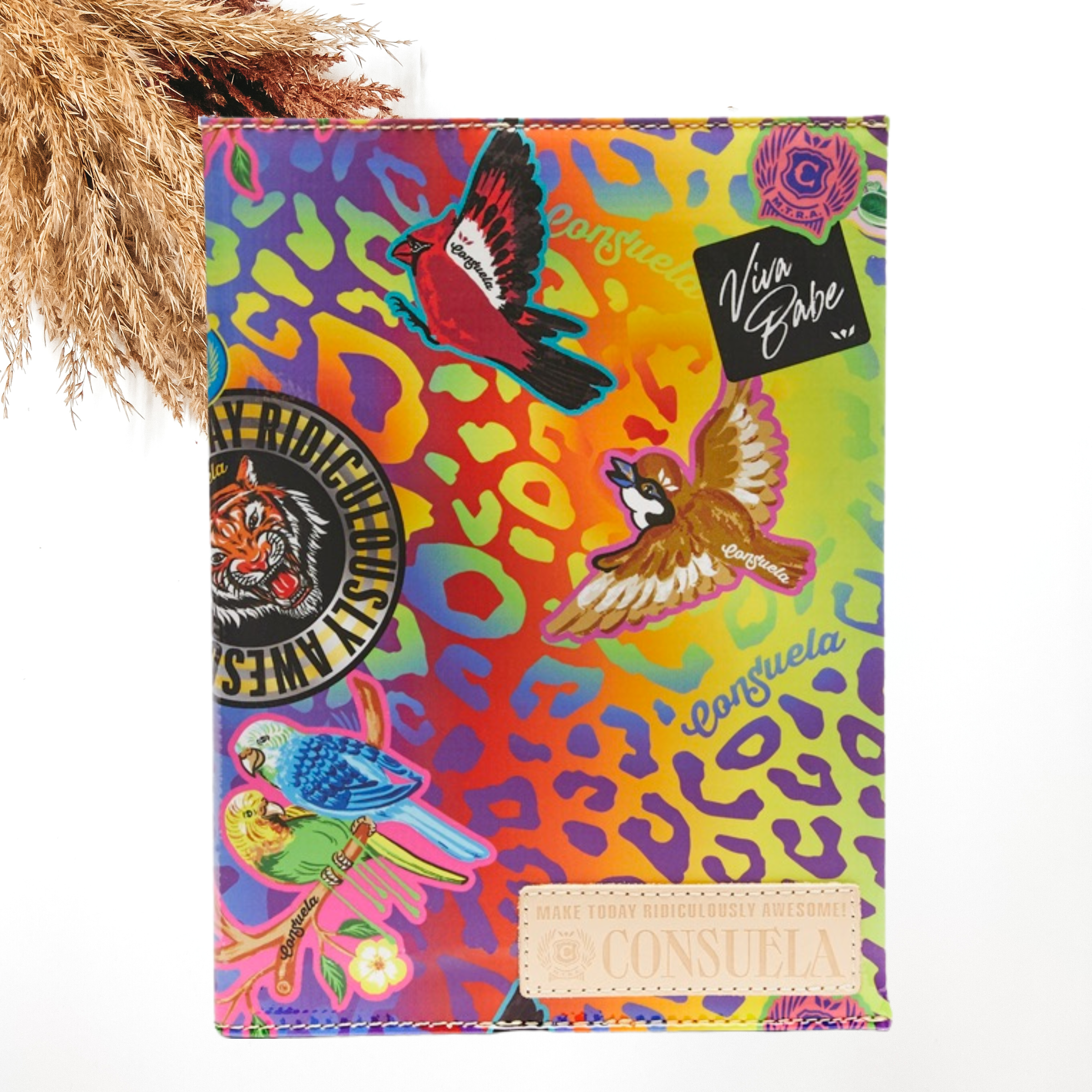 Pictured is a notebook cover with a mostly a multicolored leopard print design. It also includes random sticker patches design and a light tan Consuela patch on the bottom right corner. This notebook is pictured on a white background with pompous grass in the top left corner of the picture.
