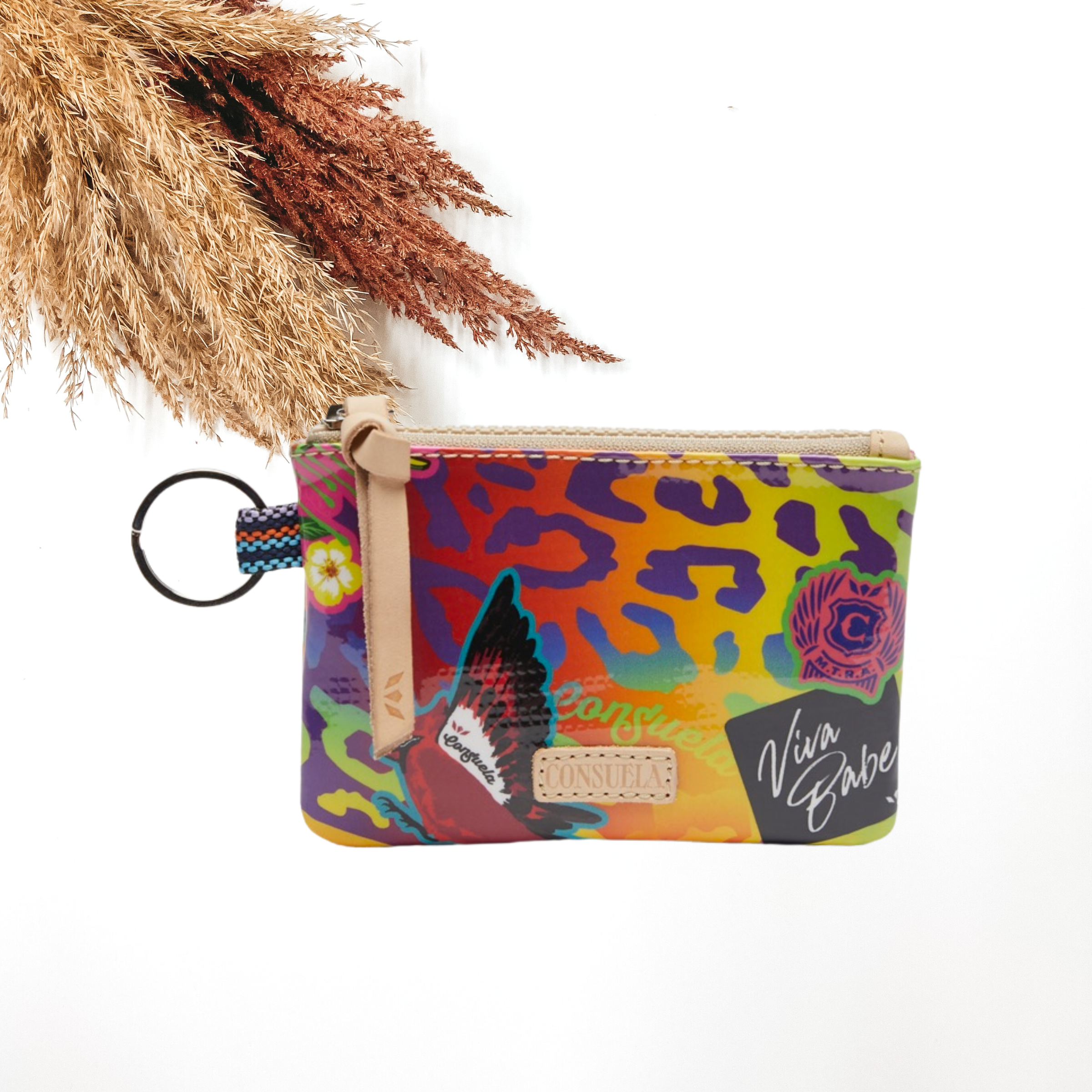 Small, rectangle pouch with a silver key ring on the left side and a light tan zipper pull. This pouch has a multicolored leopard print design with random sticker patches throughout. This purse is pictured on a white background with tan and brown pompous grass on the right side.