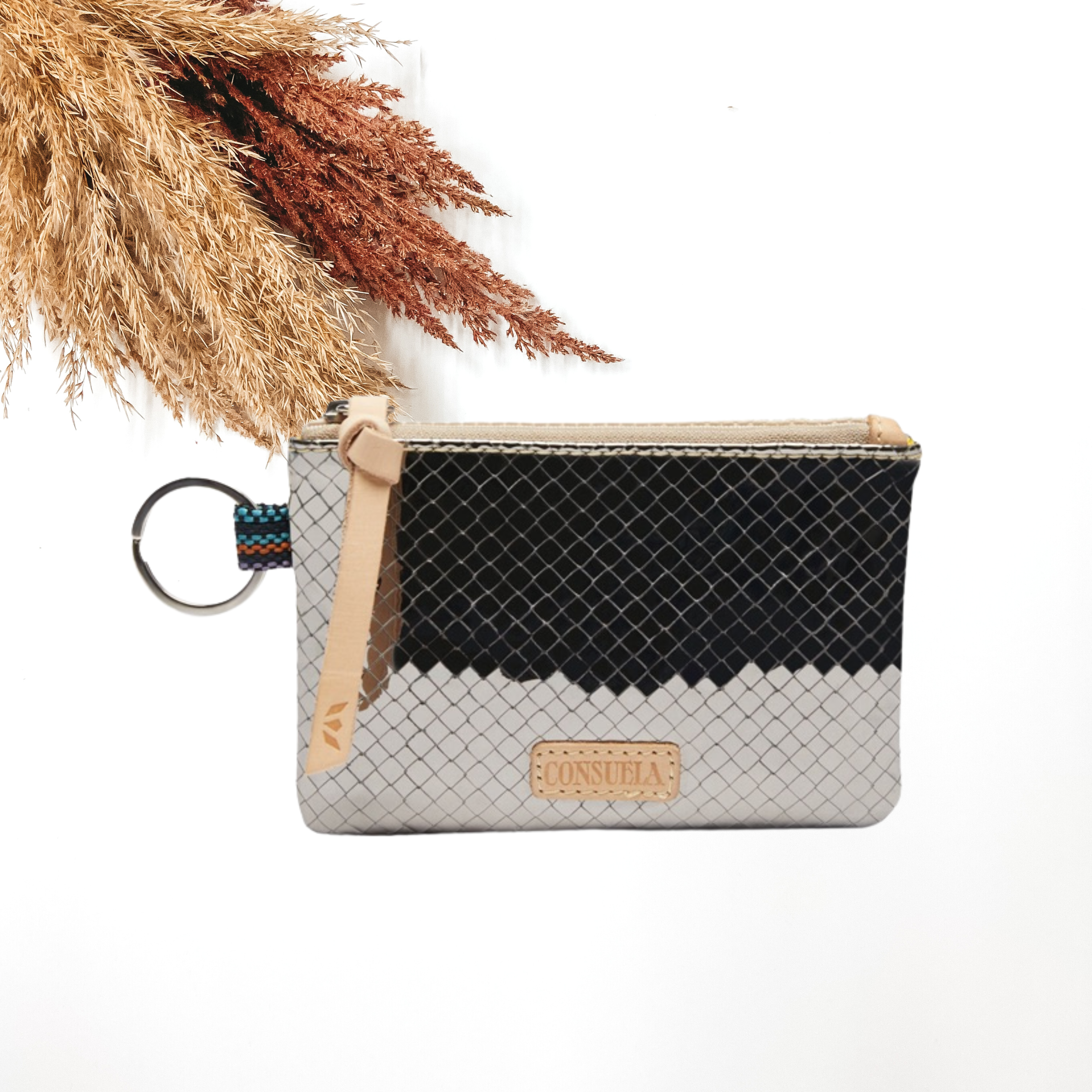 Small, rectangle pouch with a silver key ring on the left side and a light tan zipper pull. This pouch has a metallic silver color and woven print design. This purse is pictured on a white background with tan and brown pompous grass on the right side.