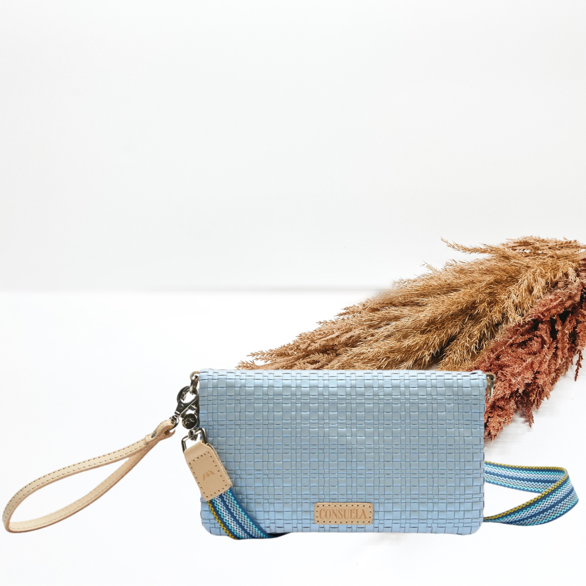 Light blue metallic rectangle purse with a woven pattern. This purse has a light tan wristlet strap and a striped, long purse strap. This pictured on a white background with tan and brown pompous grass in the background.