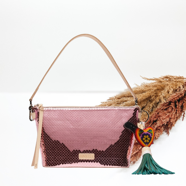 Metallic pink purse with a woven pattern. This purse includes a light tan purse strap and a heart charm with green tassels. This purse is pictured on a white background with pompous grass in the background. 