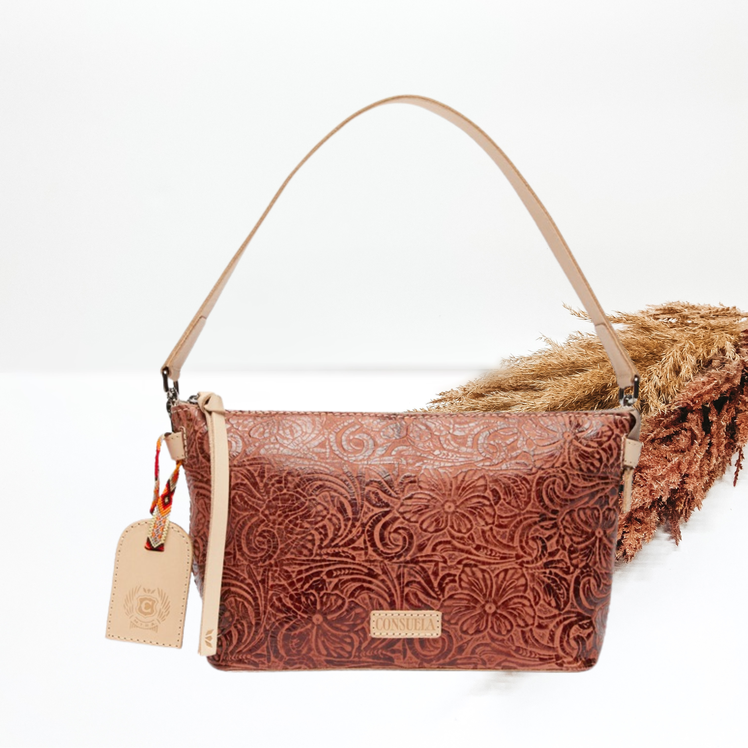 Brown leather tooled print purse. This purse includes a light tan purse strap and a light tan luggage tag charm. This purse is pictured on a white background with pompous grass in the background. 