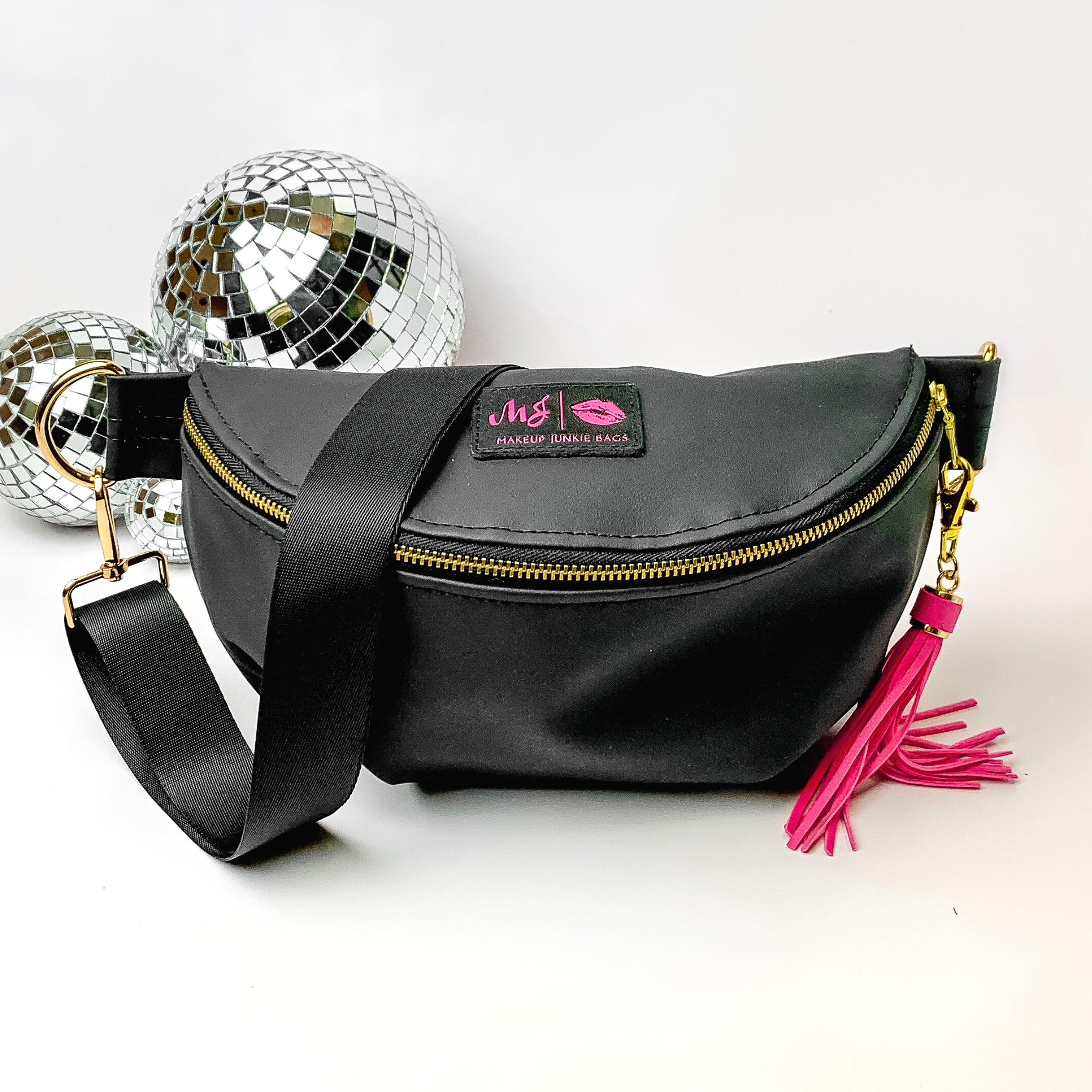 Black fanny pack with a black strap. This bag includes gold accents, a gold zipper, and a hot pink tassel. This bag also has a pink stitched logo for Makeup Junkie. This fanny pack is pictured on a white background in front of disco balls. 