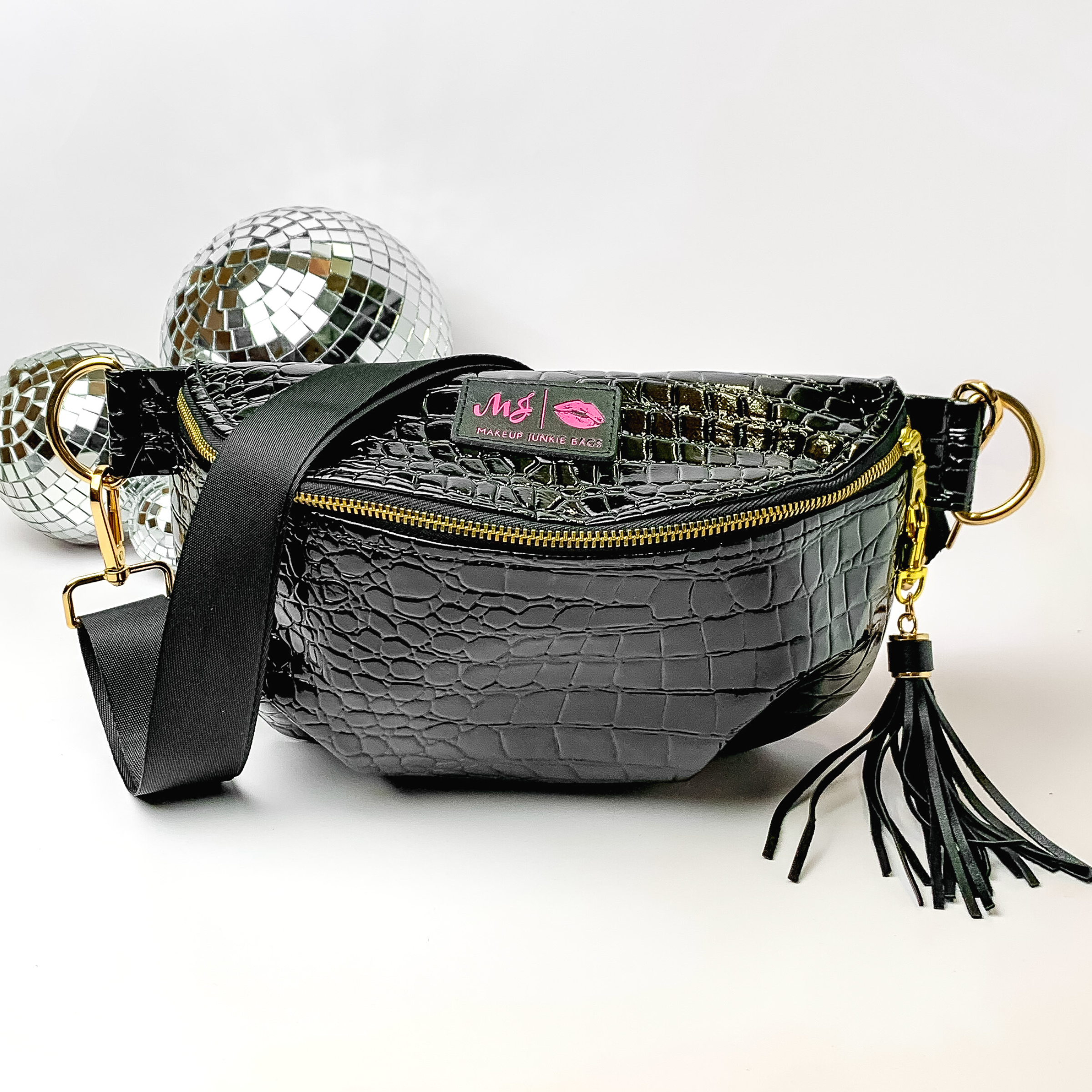 Black, croc print fanny pack with a black strap. This bag includes gold accents, a gold zipper, and a black tassel. This bag also has a pink stitched logo for Makeup Junkie. This fanny pack is pictured on a white background in front of disco balls. 