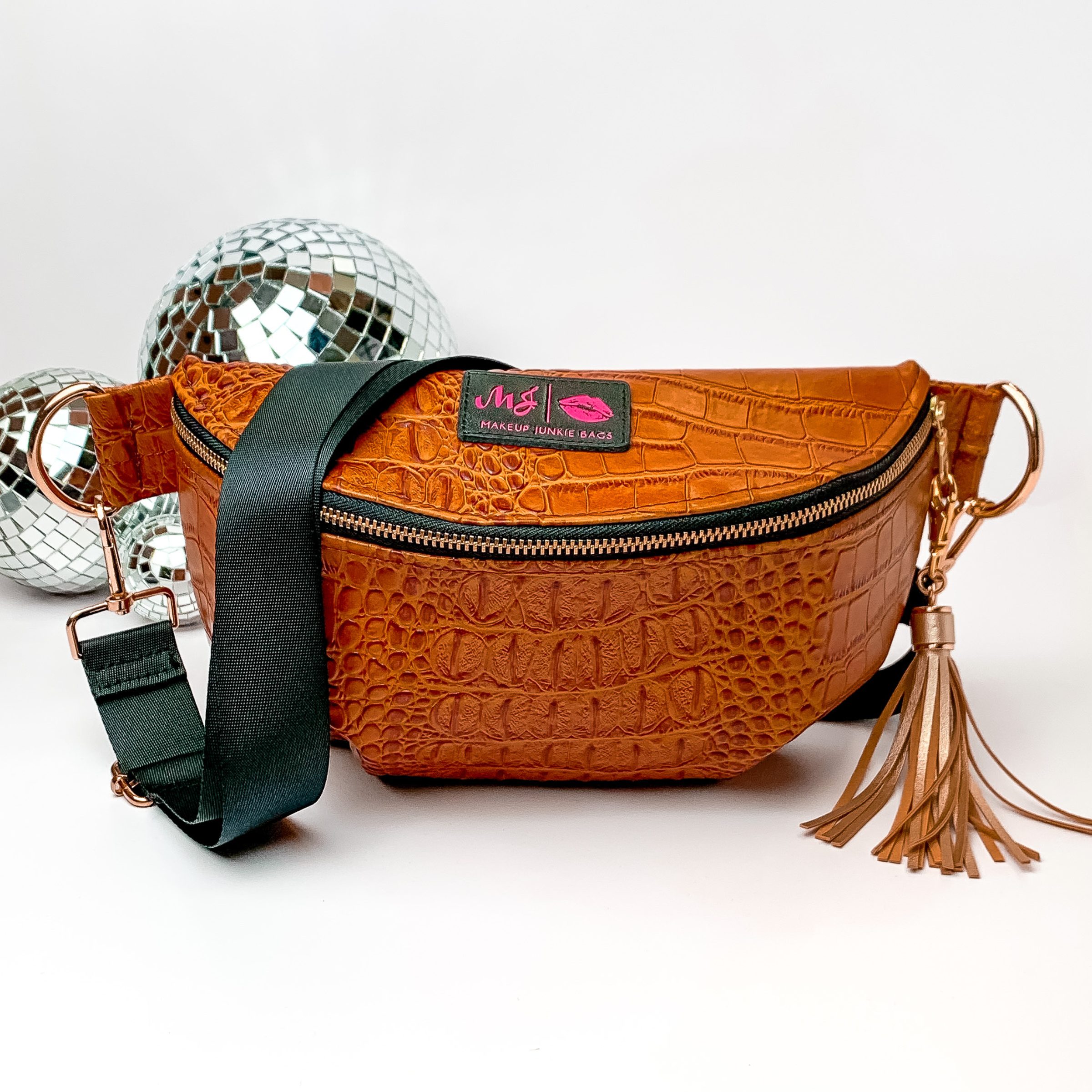 Tan, croc print fanny pack with a black strap. This bag includes gold accents, a gold zipper, and a gold tassel. This bag also has a pink stitched logo for Makeup Junkie. This fanny pack is pictured on a white background in front of disco balls. 