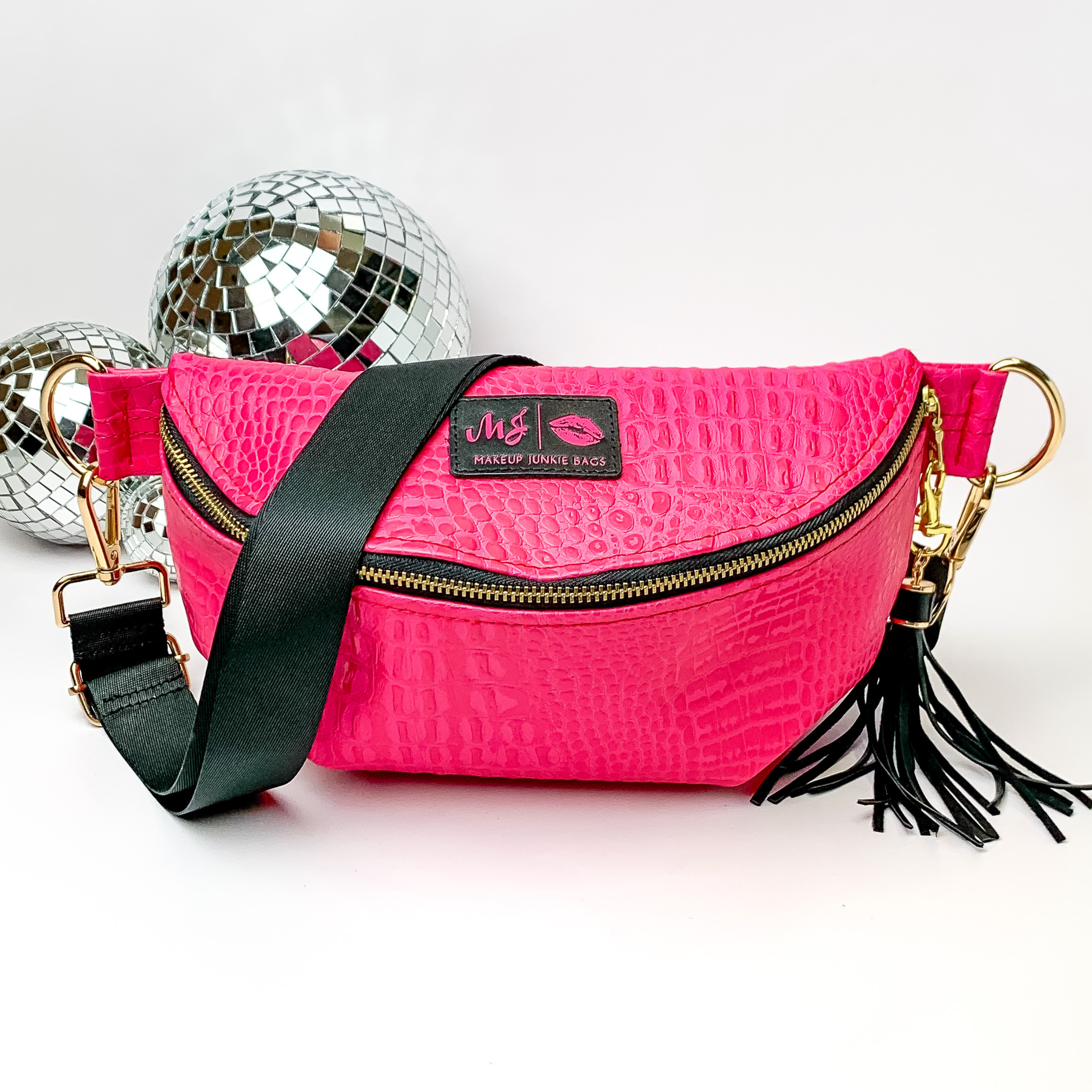 Hot pink, croc print fanny pack with a black strap. This bag includes gold accents, a gold zipper, and a gold tassel. This bag also has a pink stitched logo for Makeup Junkie. This fanny pack is pictured on a white background in front of disco balls. 