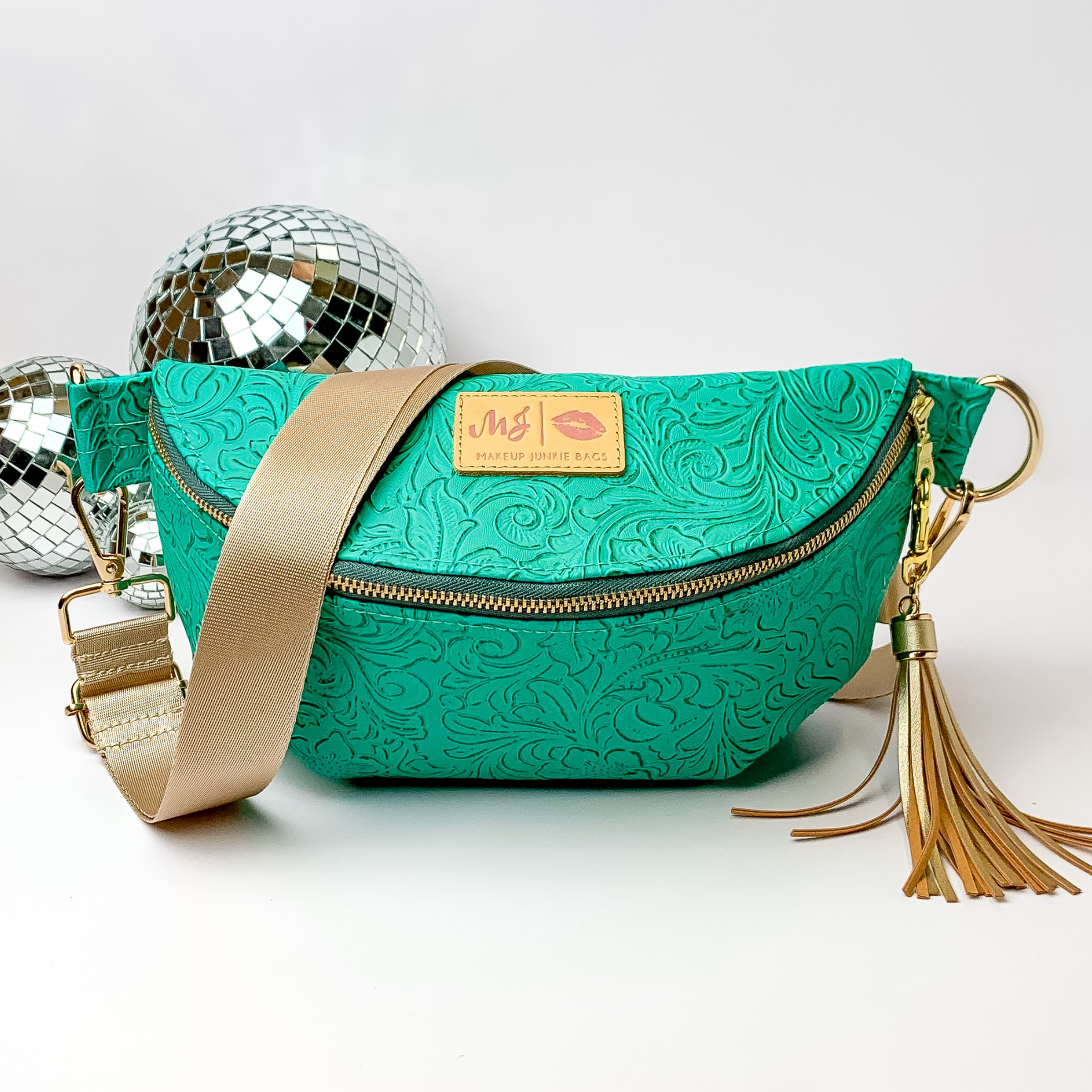 Turquoise, tooled print fanny pack with a black strap. This bag includes gold accents, a gold zipper, and a gold tassel. This bag also has a pink stitched logo for Makeup Junkie. This fanny pack is pictured on a white background in front of disco balls. 