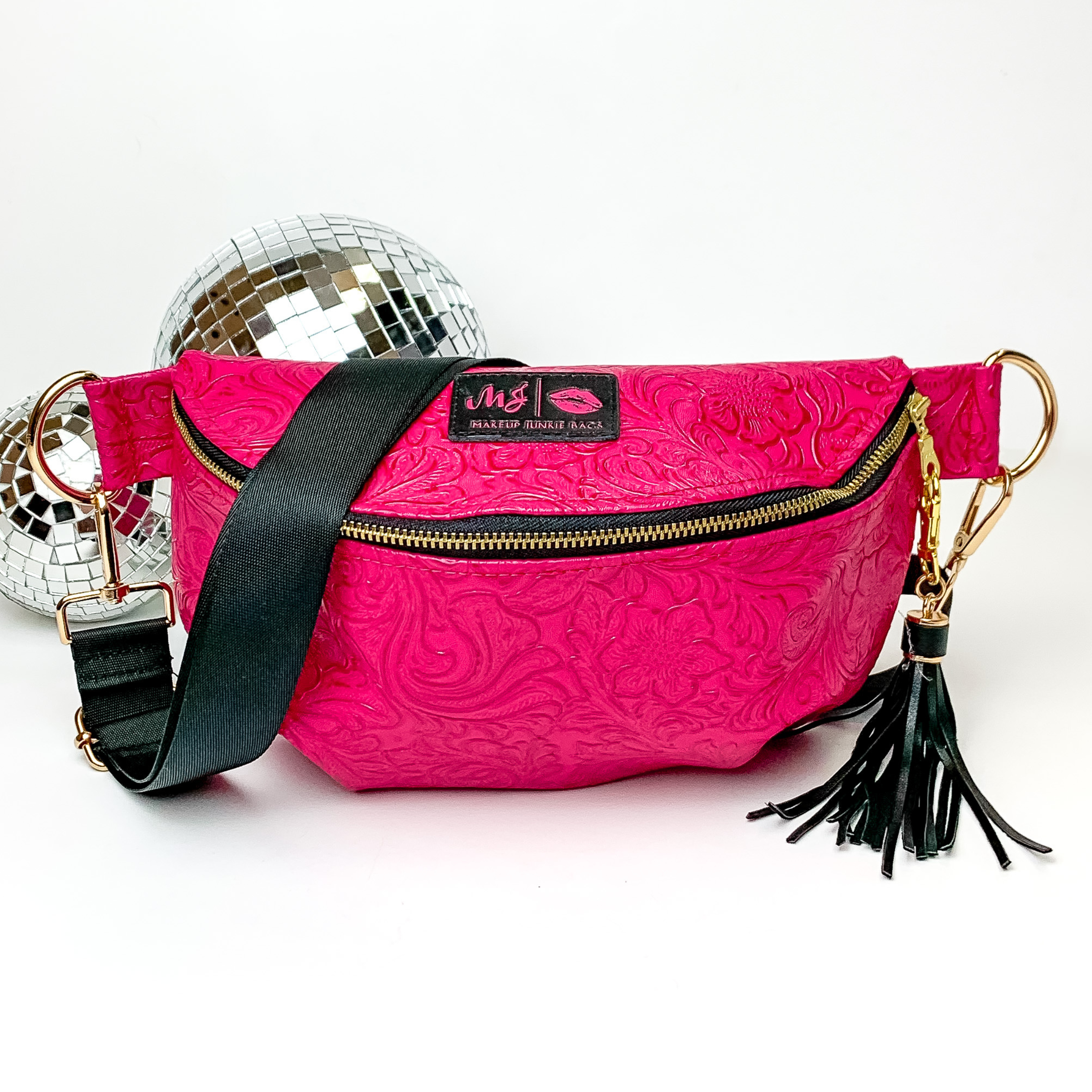 Hot pink, tooled print fanny pack with a black strap. This bag includes gold accents, a gold zipper, and a gold tassel. This bag also has a pink stitched logo for Makeup Junkie. This fanny pack is pictured on a white background in front of disco balls. 