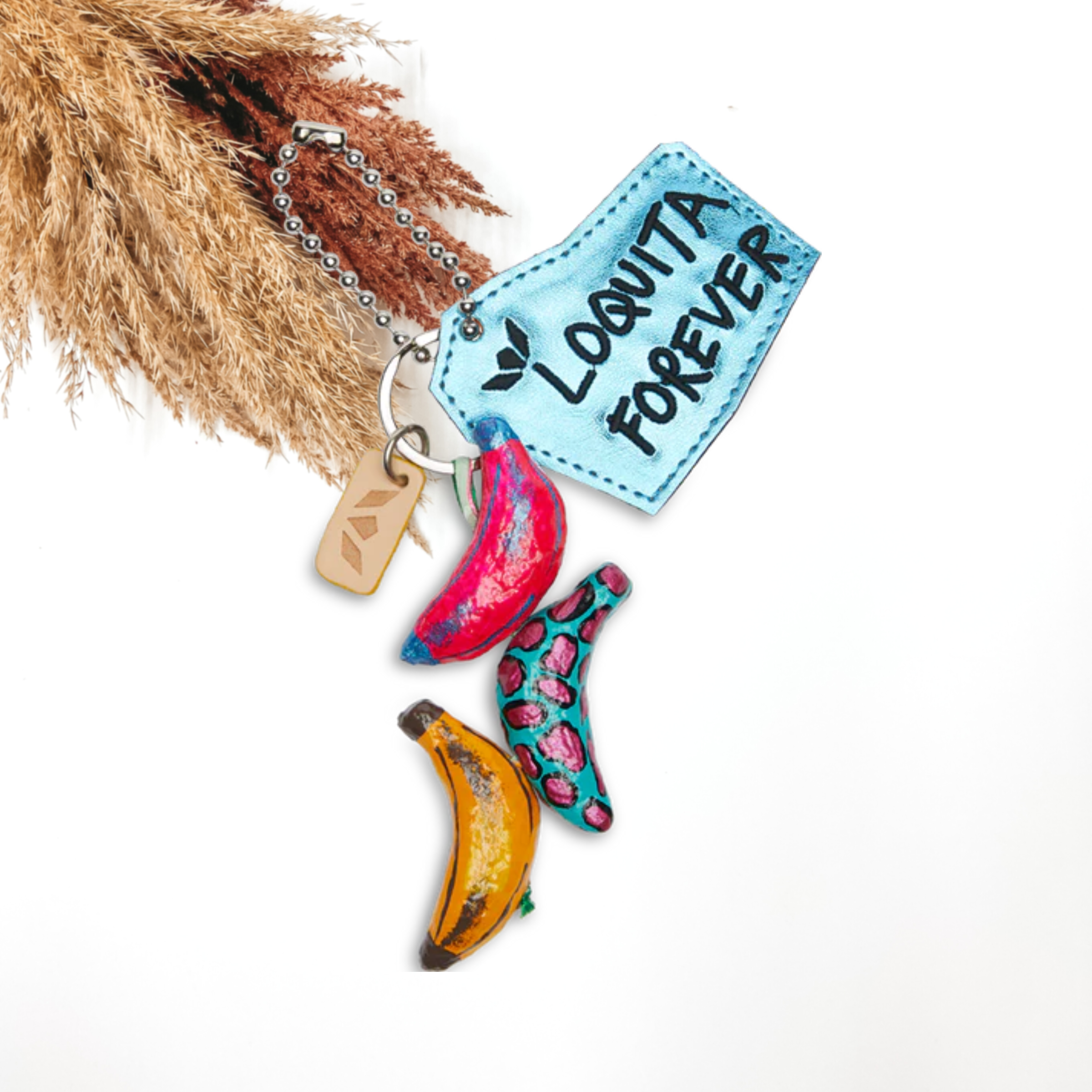 Silver key chain with hanging, colorful bananas and an irregular shaped metallic blue charm. the bananas are multicolored with different designs and the blue charm has the words "LOGUITA FOREVER" stitched in black. This charm is pictured on a white background with pompous grass at the top left corner. 