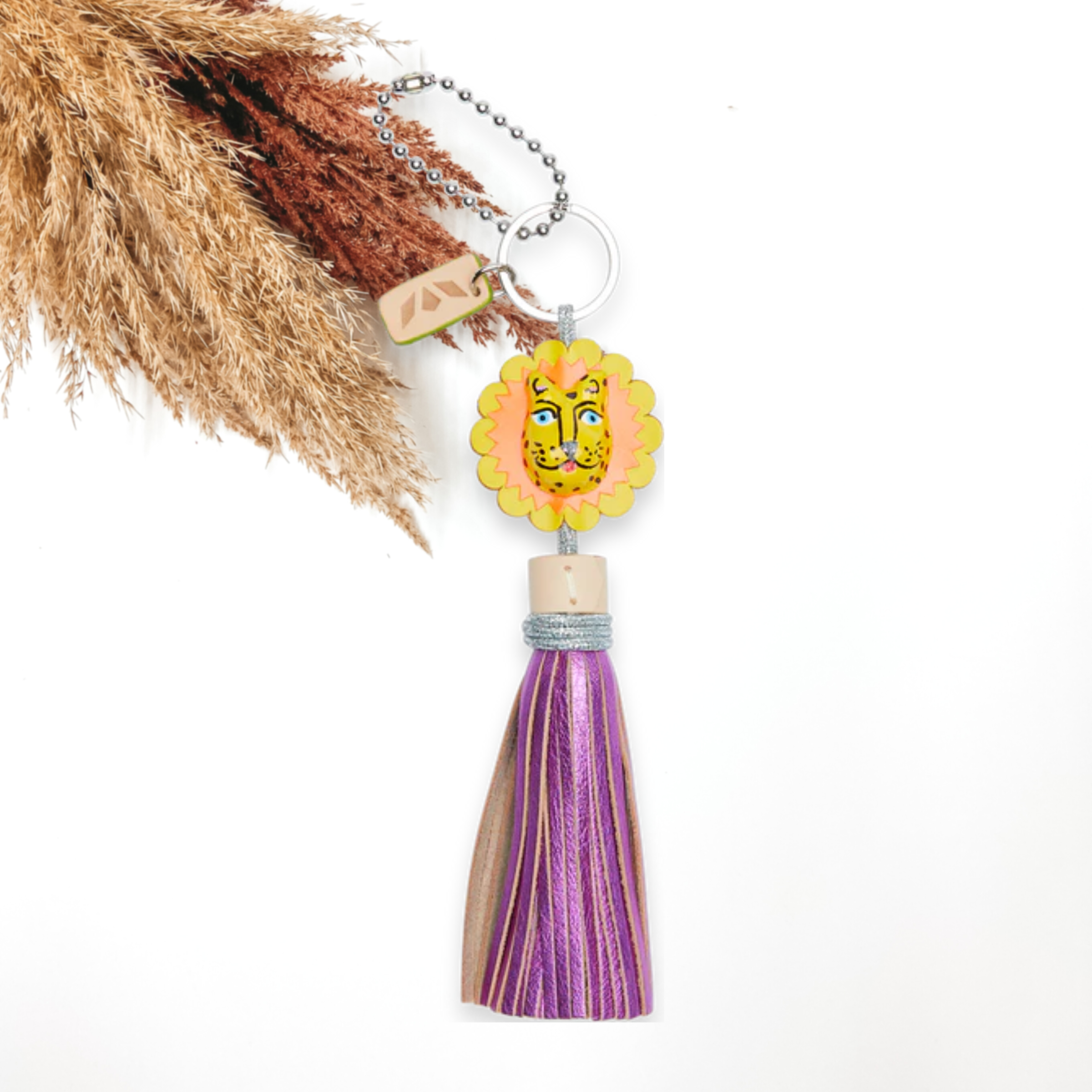 Silver key chain with a lion head charm that has a metallic purple tassel at the bottom of the lion head. This charm is pictured on a white background with pompous grass at the top left corner. 