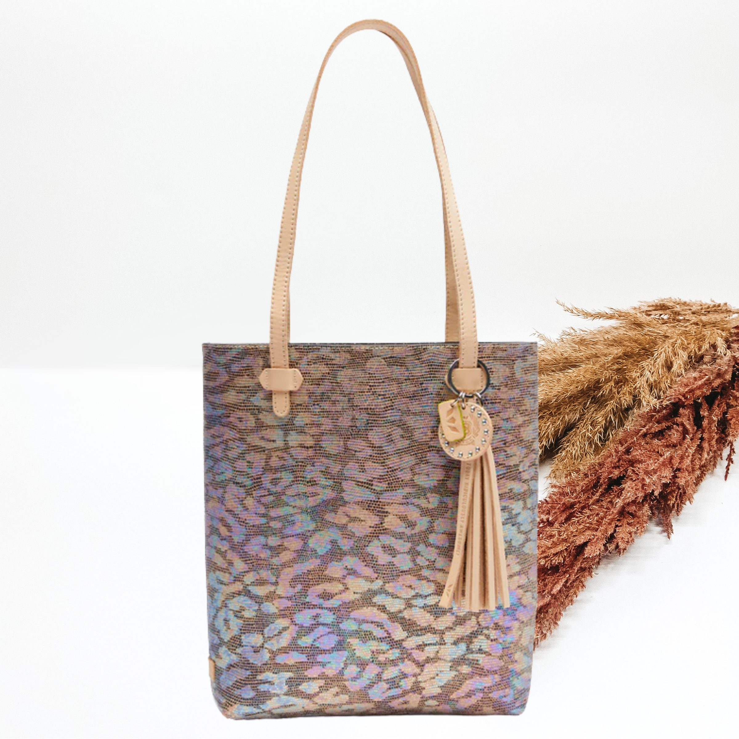 Pictured is long, rectangle tote that has a metallic, multicolored leopard print design. This purse also includes light tan, leather straps. This purse is pictured on a white background with pompous grass on the right side of the picture.
