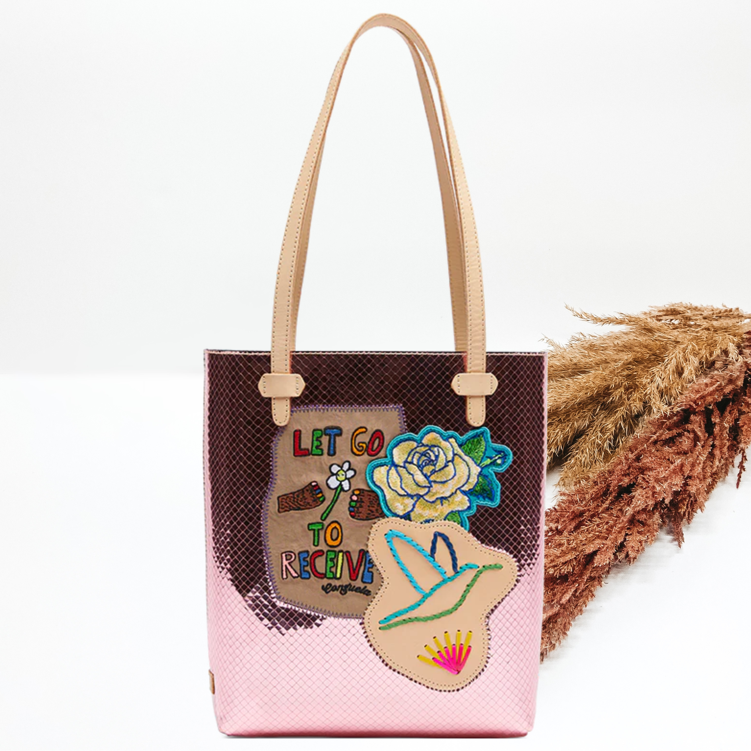 Pictured is long, rectangle tote that has a metallic pink color with a woven design ith light tan straps. This purse also a rose patch, and tan leather patch with a stitched bird, and a patch with the words "LET GO TO RECEIVE". This purse is pictured on a white background with pompous grass on the right side of the picture.
