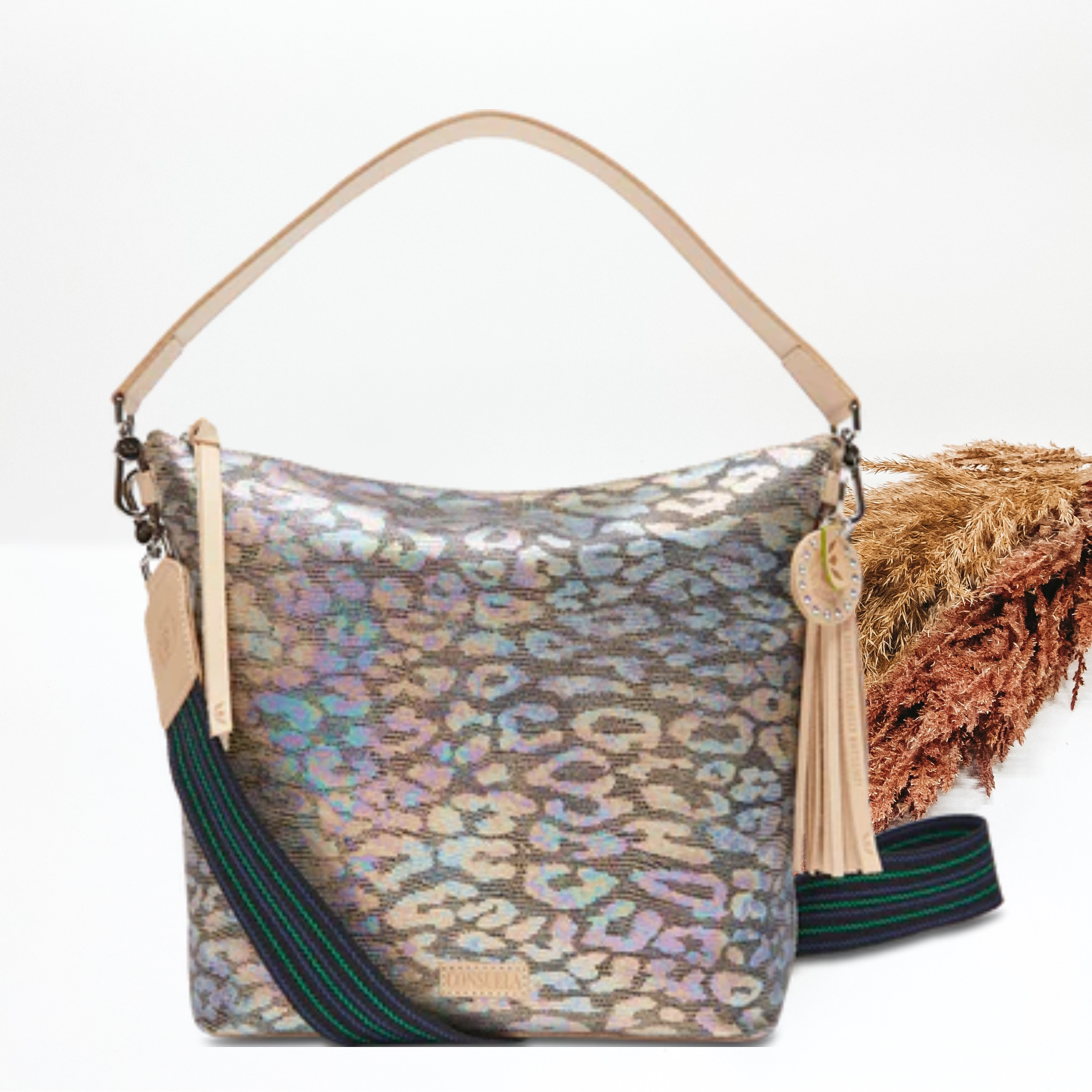 Pictured is hobo purse that has a metallic, multicolor leopard print deisgn. This purse includes a thick, light tan strap and striped, thick purse strap. This purse is pictured on a white background with pompous grass on the right side of the picture.