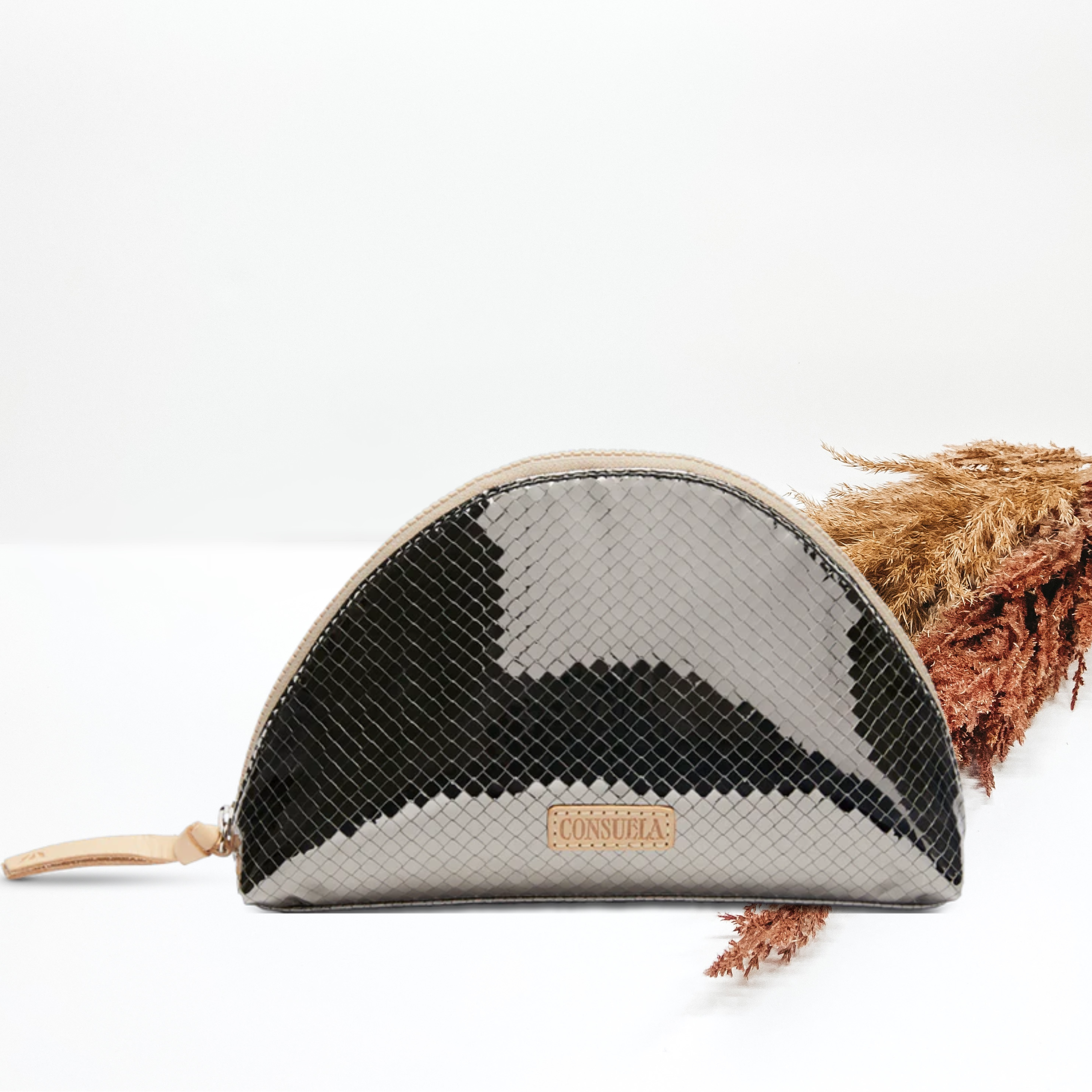Pictured is a dome shaped cosmetic bag. This bag is metallic silver with a woven design and includes a top zipper. This purse is pictured on a white background with pompous grass on the right side of the picture.