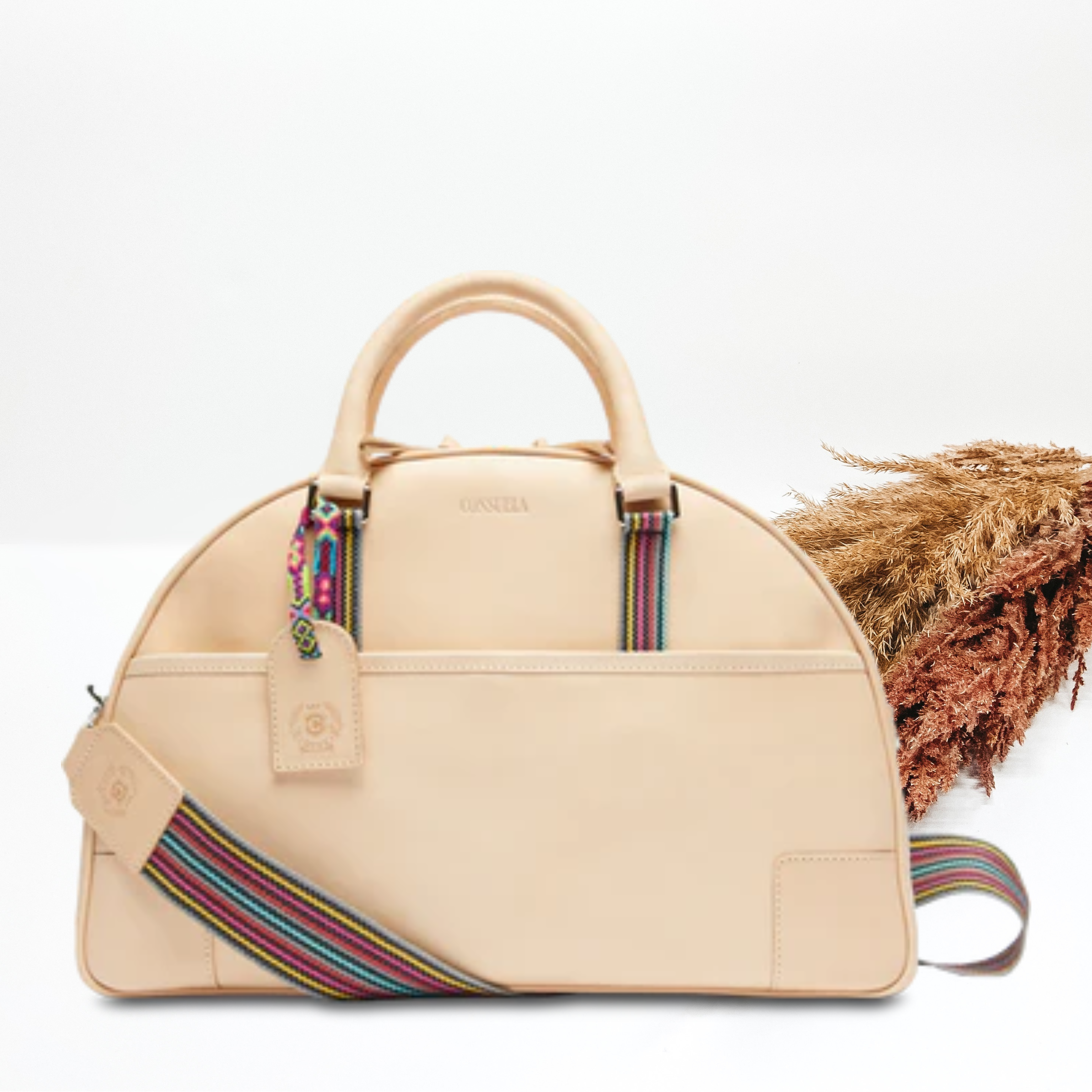 Pictured is a dome shaped bag with light tan short handles and a striped strap. This bag is a light tan leather with light tan accents and a charm on one of the handles. This bag is pictured on a white background with pompous grass on the right side of the bag.