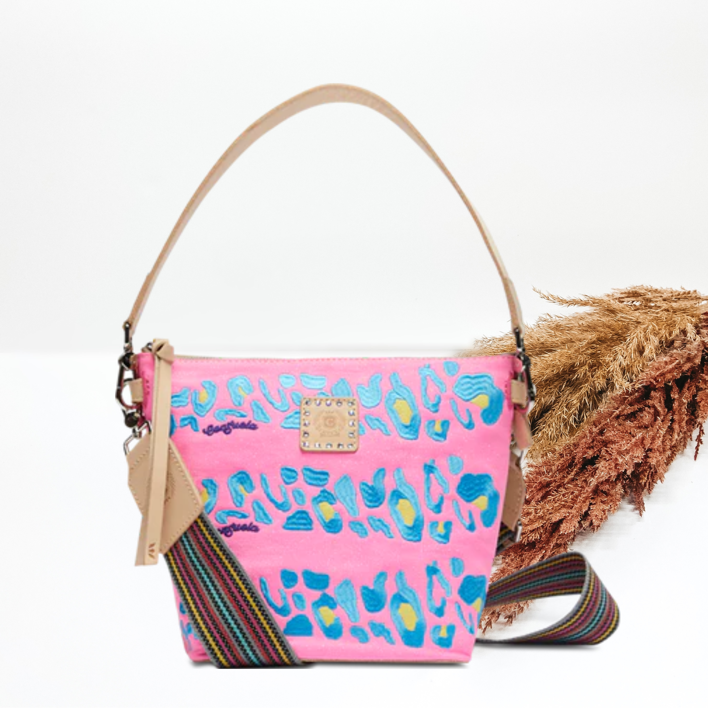 Pictured is a wedge purse in light pink with a light blue and yellow stiched leopard print pattern. This purse has a light tan short strap with a thick, striped longer purse strap This bag is pictured on a white background with pompous grass on the left side of the picture. 