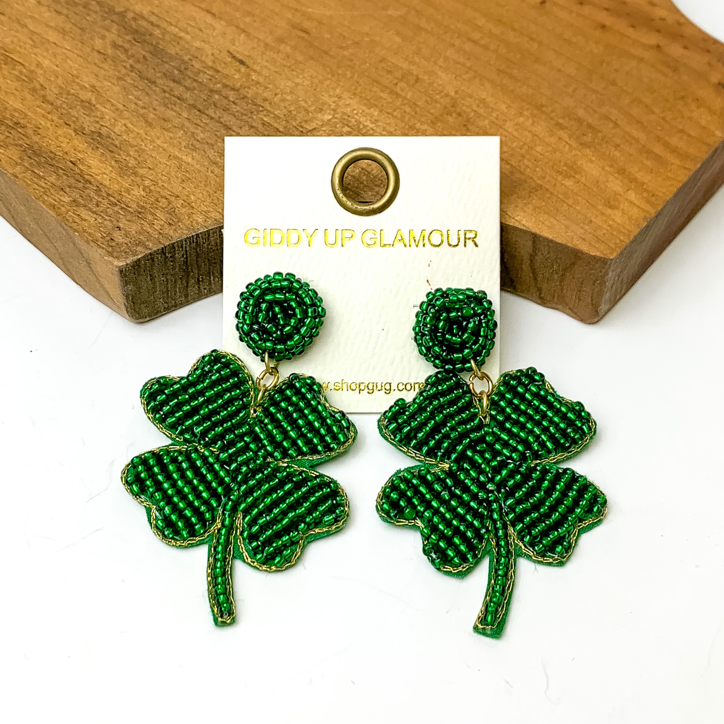 Green beaded circle post earrings with a green beaded four leaf clover drop earrings. These earrings are pictured in front of a brown block on a white background. 