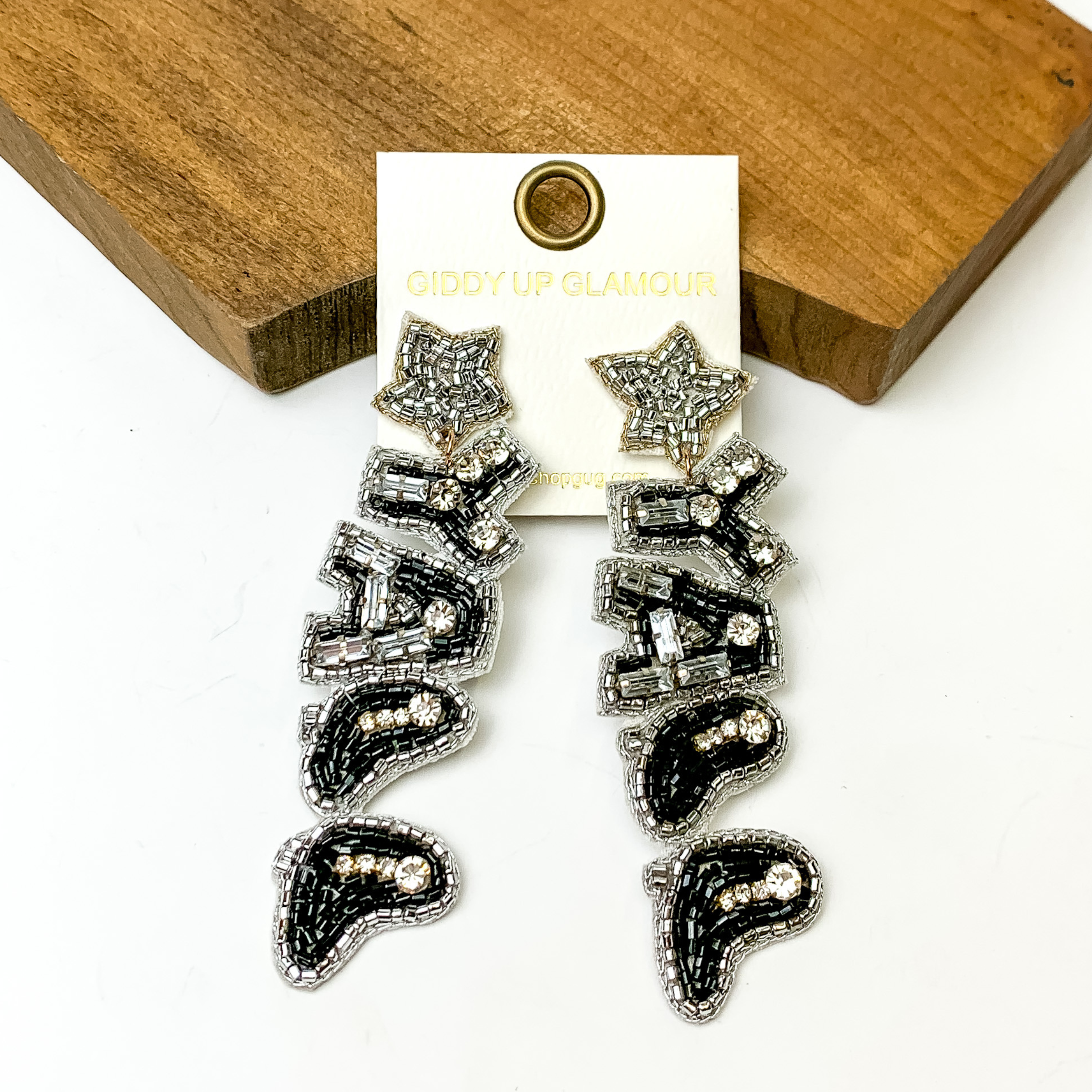 Silver beaded star post back earrings with a hanging pendant that spells out "YALL". The word is mosty black beads outlines in silver and clear rystals throughout. The two "L"s are in the shape of boots. These earrings are pictured on a white background with a brown block at the top of the picture.