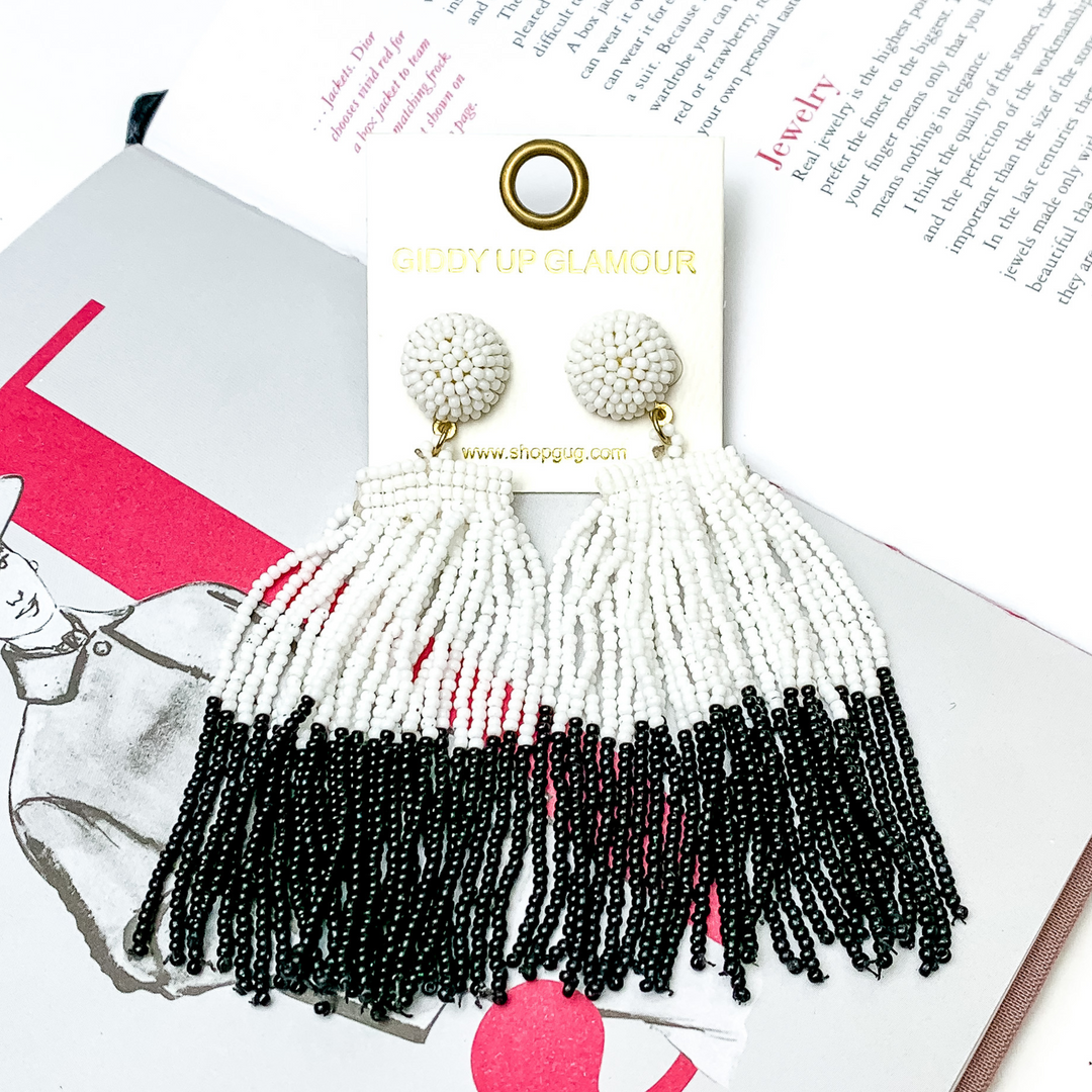 These are circle post back, beaded earrings in turquoise with a hanging beaded tassel. The tassel is half white and half black. These earrings are pictured on top of an open book on a white background. 