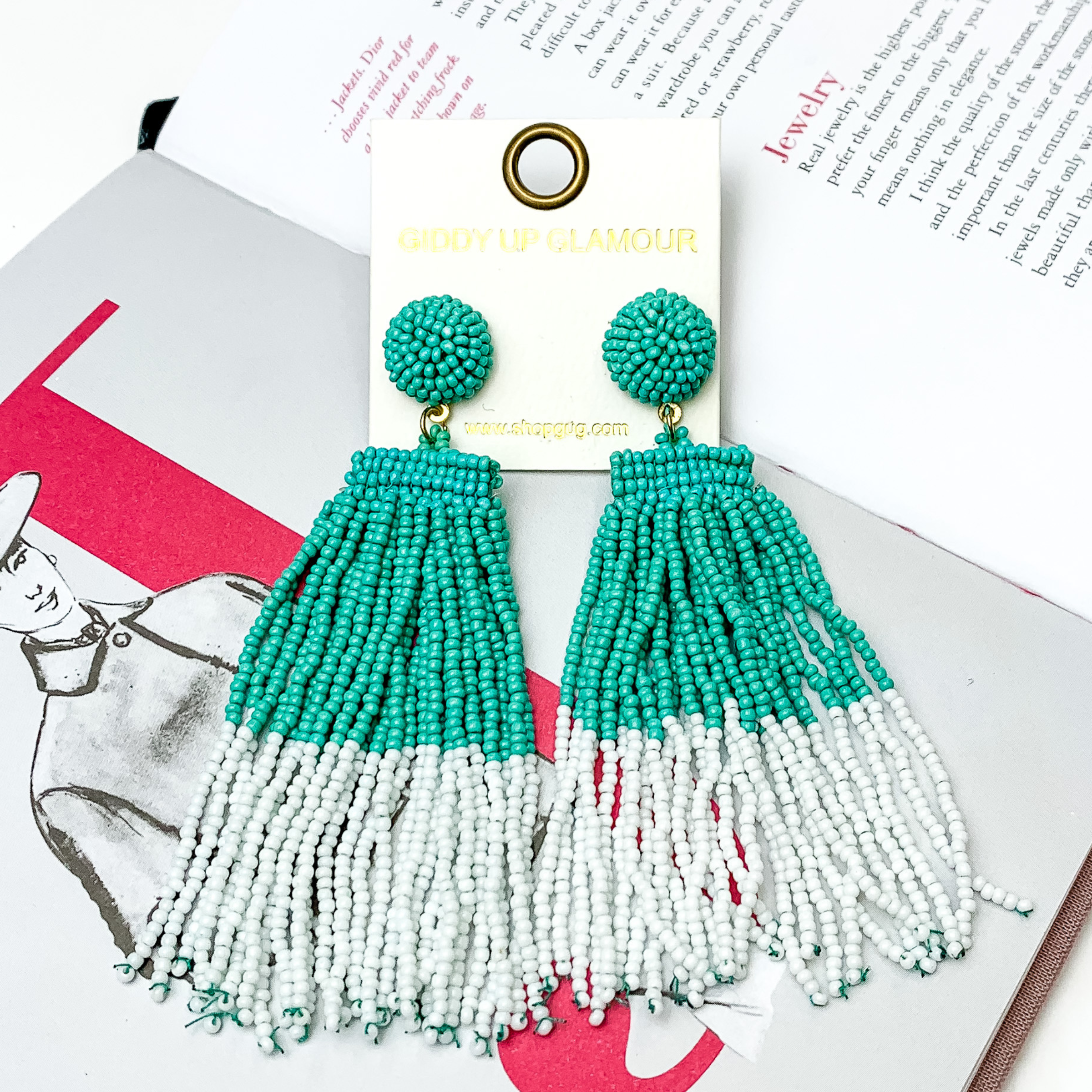 These are circle post back, beaded earrings in turquoise with a hanging beaded tassel. The tassel is half turquoise and half white. These earrings are pictured on top of an open book on a white background. 