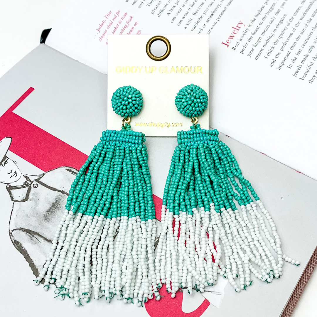 These are circle post back, beaded earrings in turquoise with a hanging beaded tassel. The tassel is half turquoise and half white. These earrings are pictured on top of an open book on a white background. 