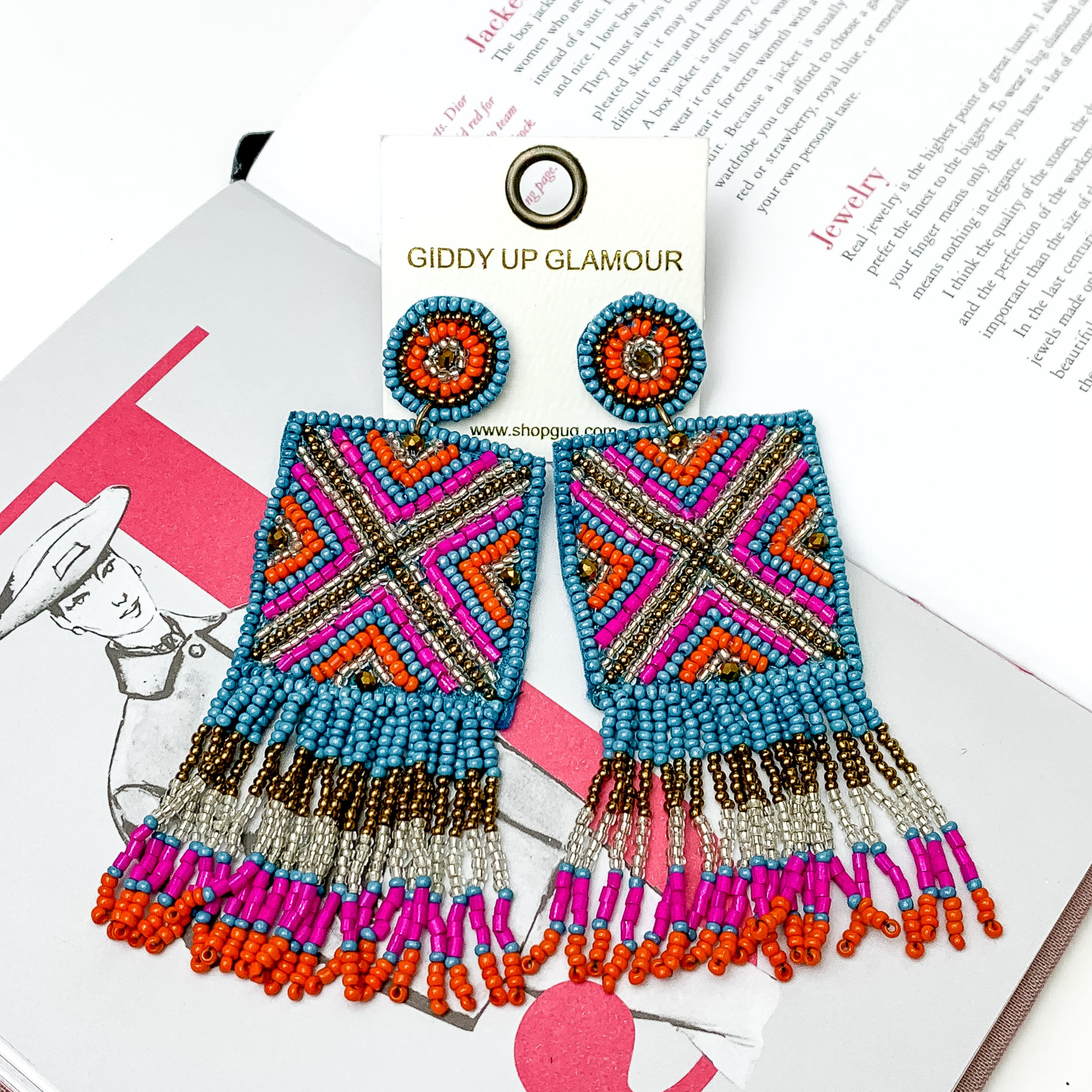 These are circle post back, beaded earrings with a beaded square drop that has fringe on the bottom side of the square. These earrings include blue beads, fuchsia pink beads, orange beads and gold beads. These earrings are pictured on top of an open book on a white background. 