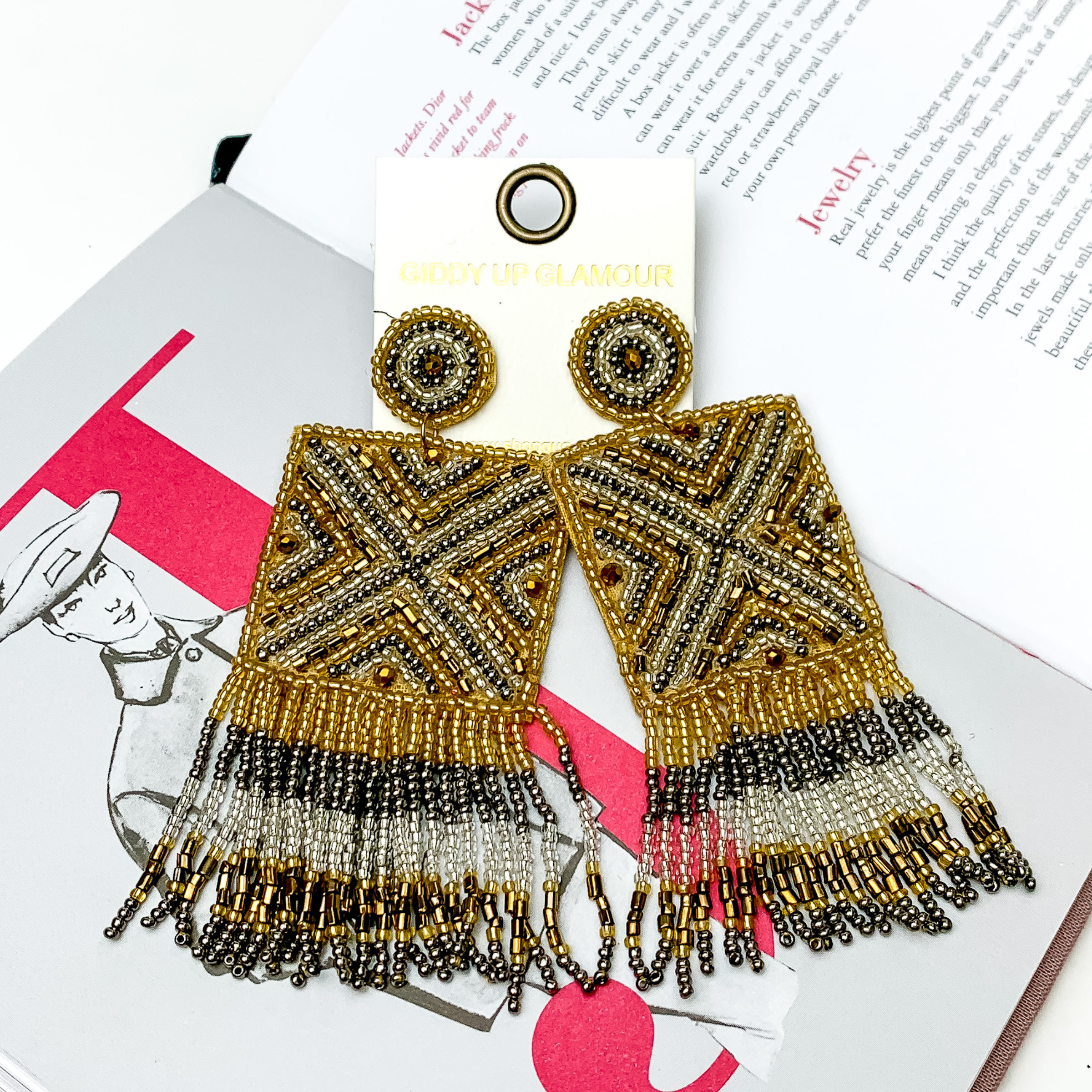 These are circle post back, beaded earrings with a beaded square drop that has fringe on the bottom side of the square. These earrings include gold beads, silver beads, and clear beads. These earrings are pictured on top of an open book on a white background. 