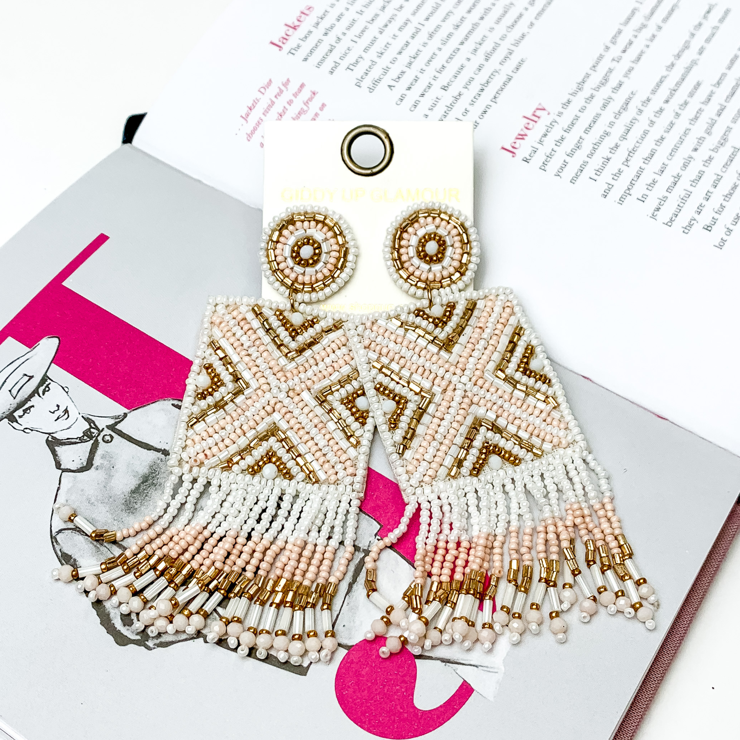 These are circle post back, beaded earrings with a beaded square drop that has fringe on the bottom side of the square. These earrings include white beads, pale pink beads, and gold beads. These earrings are pictured on top of an open book on a white background. 