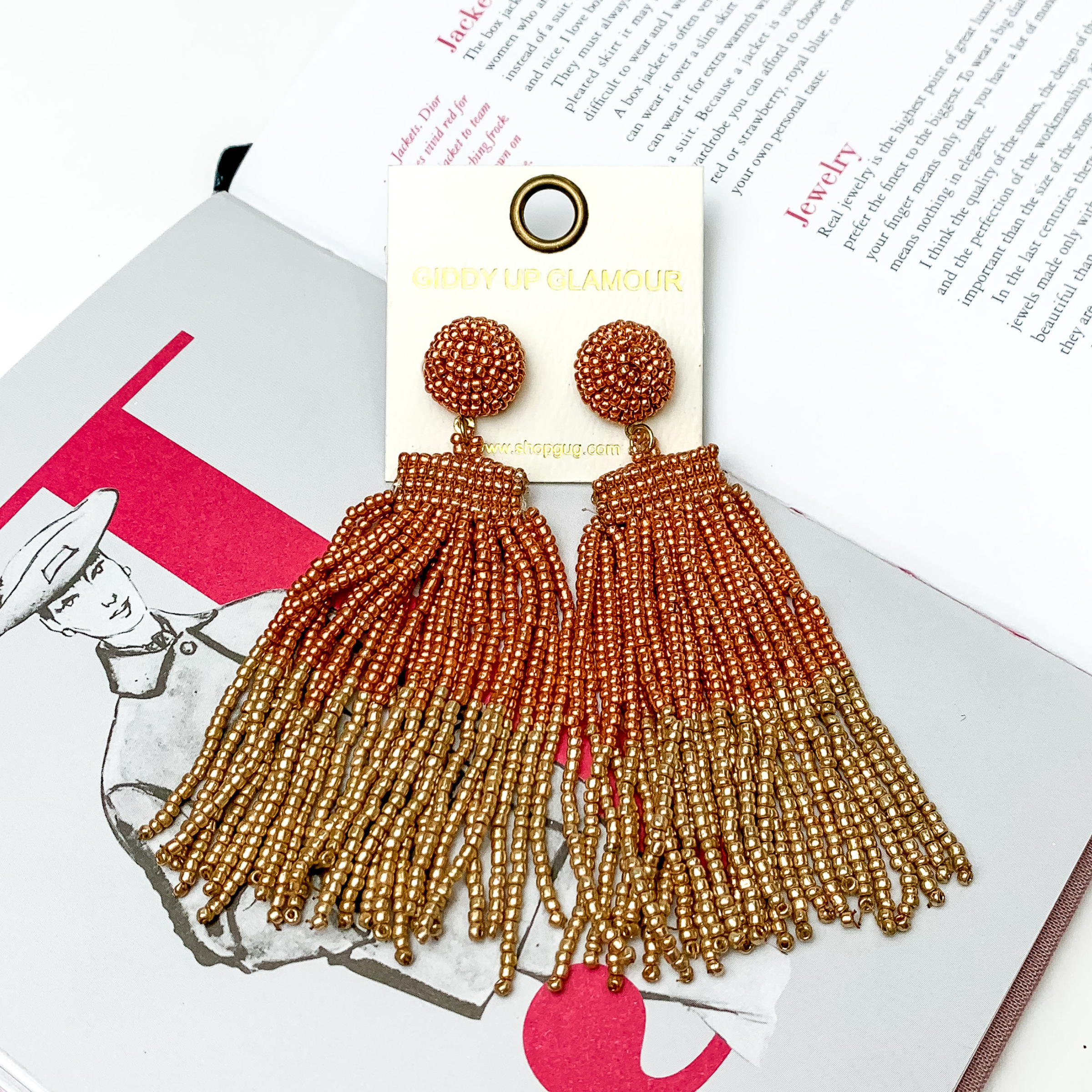 These are circle post back, beaded earrings in rose gold with a hanging beaded tassel. The tassel is half rose gold and half gold. These earrings are pictured on top of an open book on a white background. 