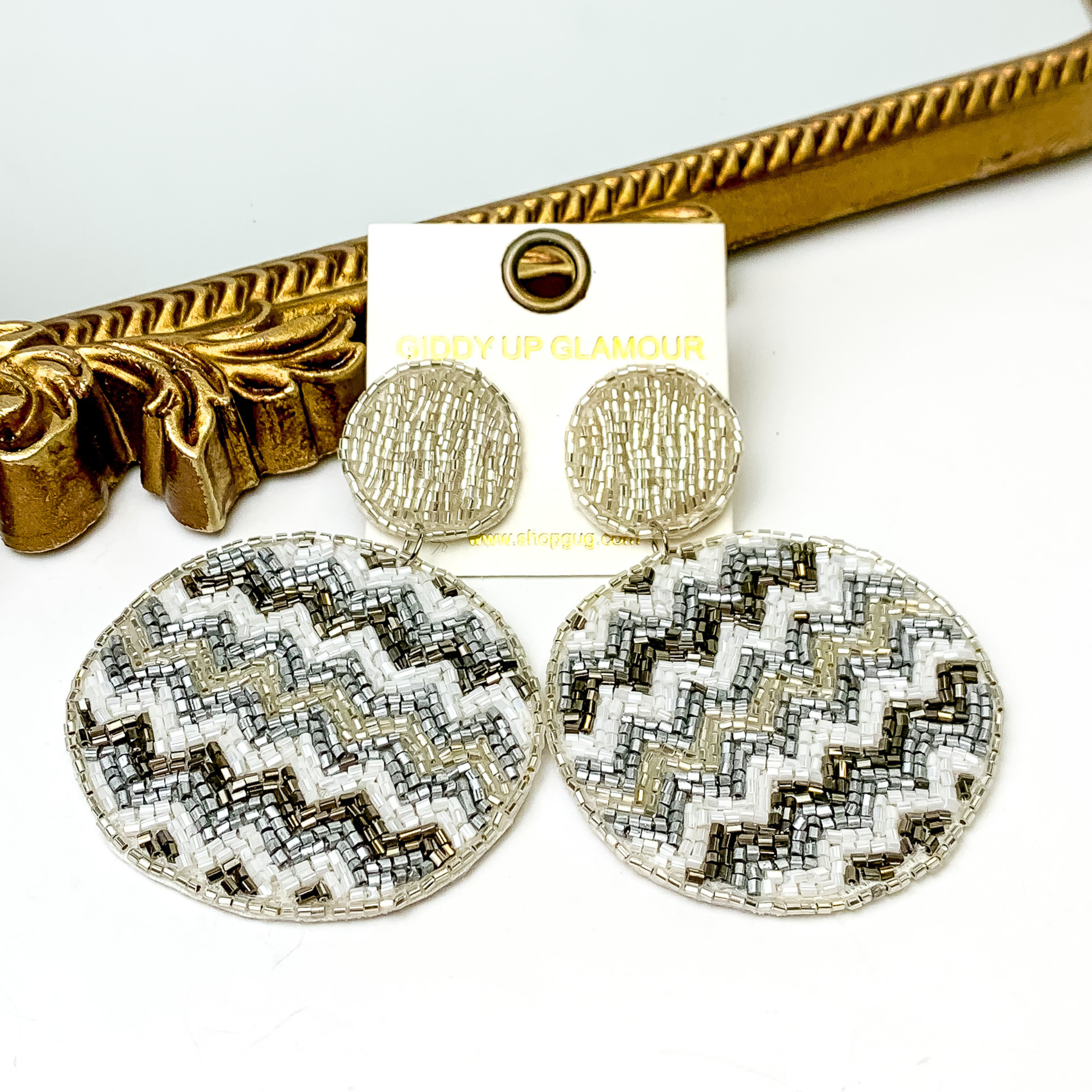 These are circle post back, light silver beaded earrings with a beaded circle drop. These earrings include a chevron pattern with white beads, silver beads, dark silver beads and light silver beads. These earrings are pictured in front of a gold mirror on a white background. 