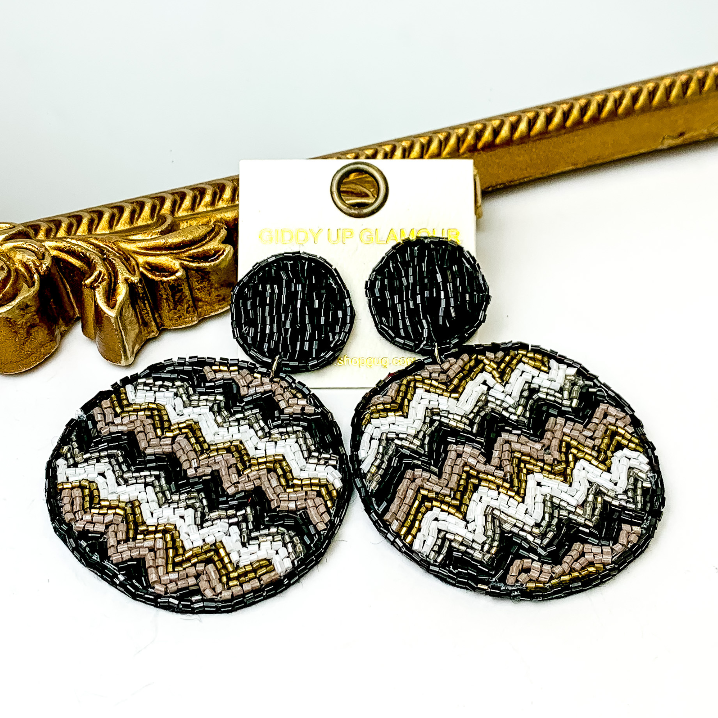 These are circle post back, black beaded earrings with a beaded circle drop. These earrings include a chevron pattern with bronze beads, silver beads,white beads and black beads. These earrings are pictured in front of a gold mirror on a white background. 
