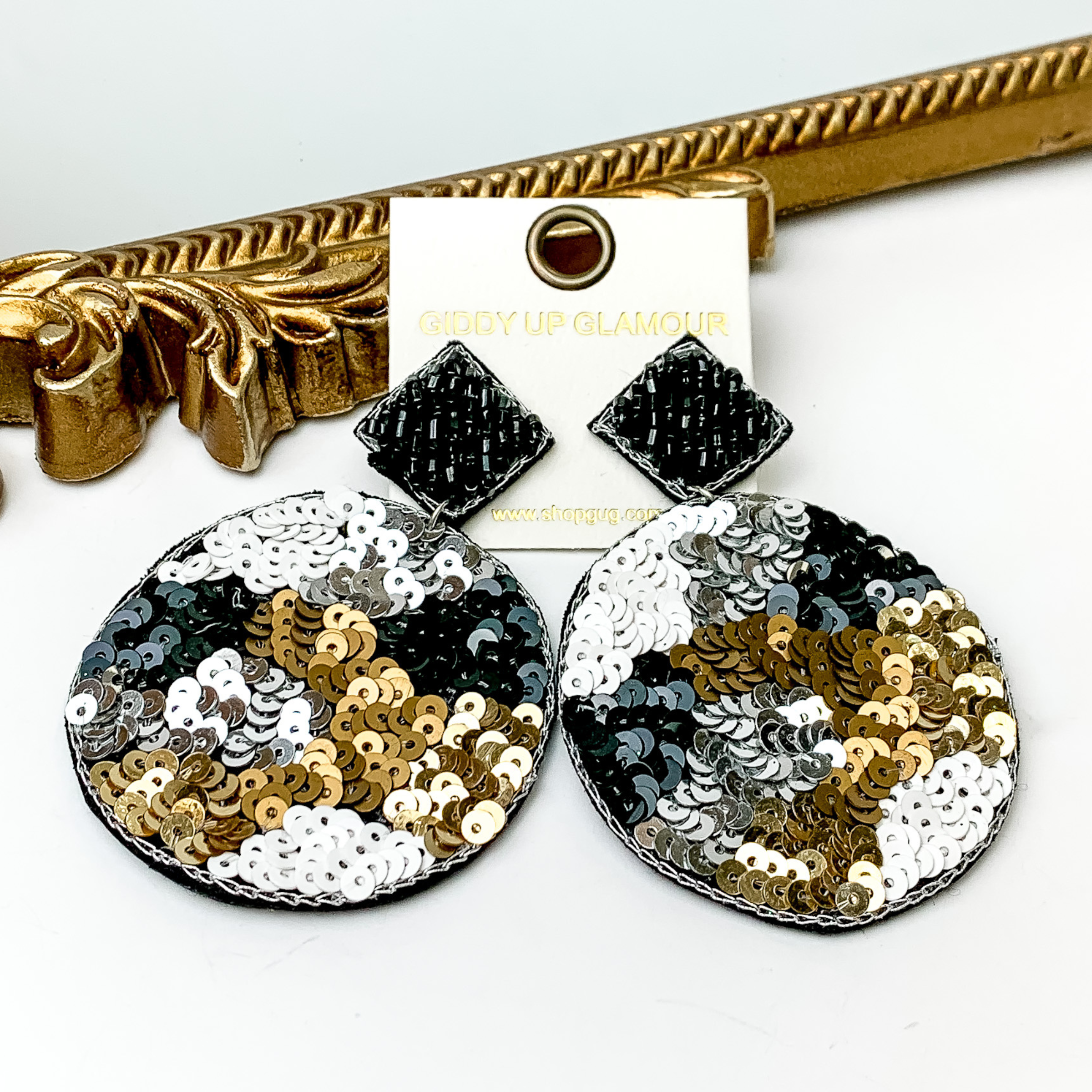 These are diamond post back, black beaded earrings with a sequin beaded circle drop. These earrings include random spot pattern with bronze sequins, white sequins, silver sequins and black sequins. These earrings are pictured in front of a gold mirror on a white background. 