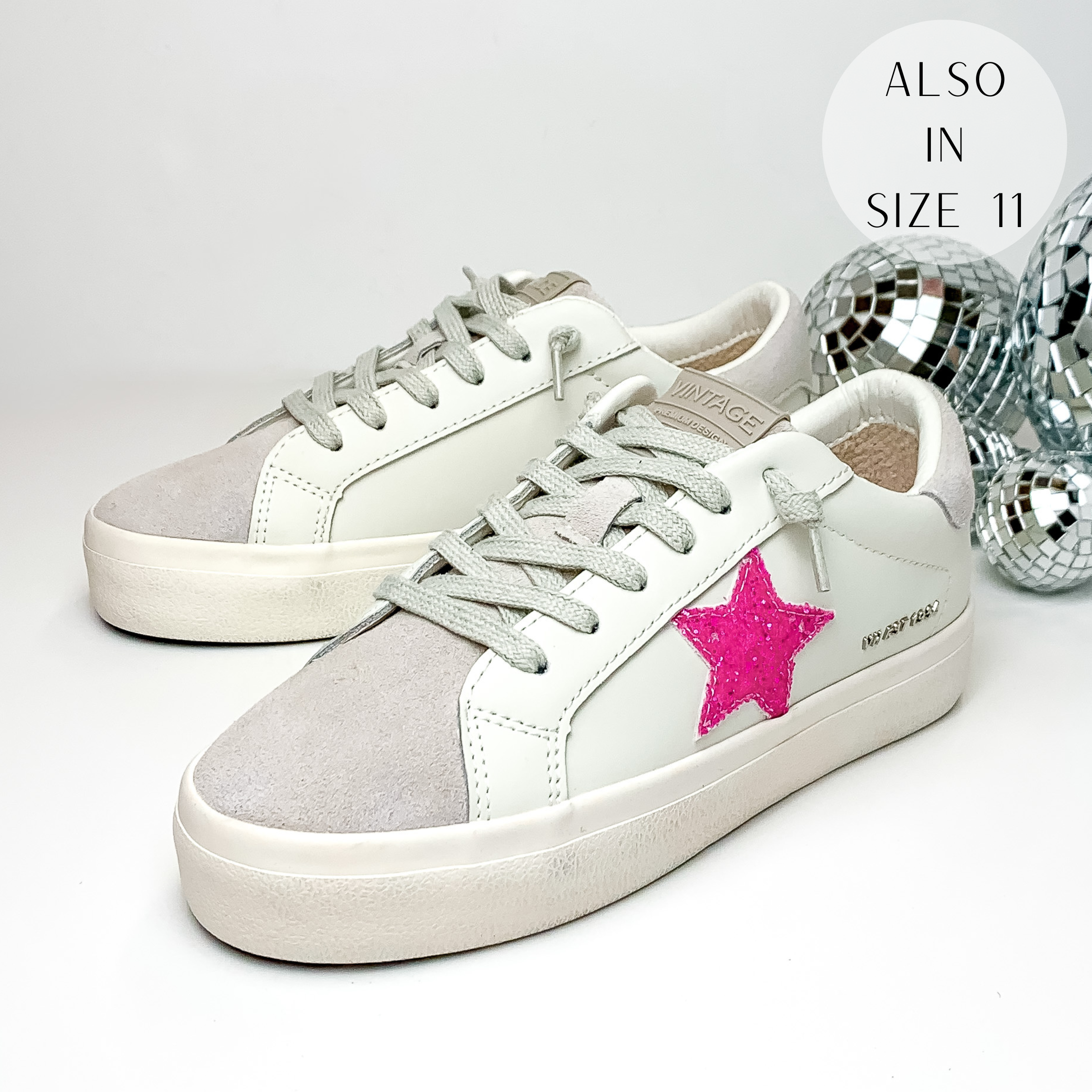 White tennis shoes with a light grey fabric on the tongue and on the back of the heel. These shoes also have a bright pink glitter star emblem on the side of the shoe. These shoes are pictured on a white background with disco balls behind them on the right hand side.