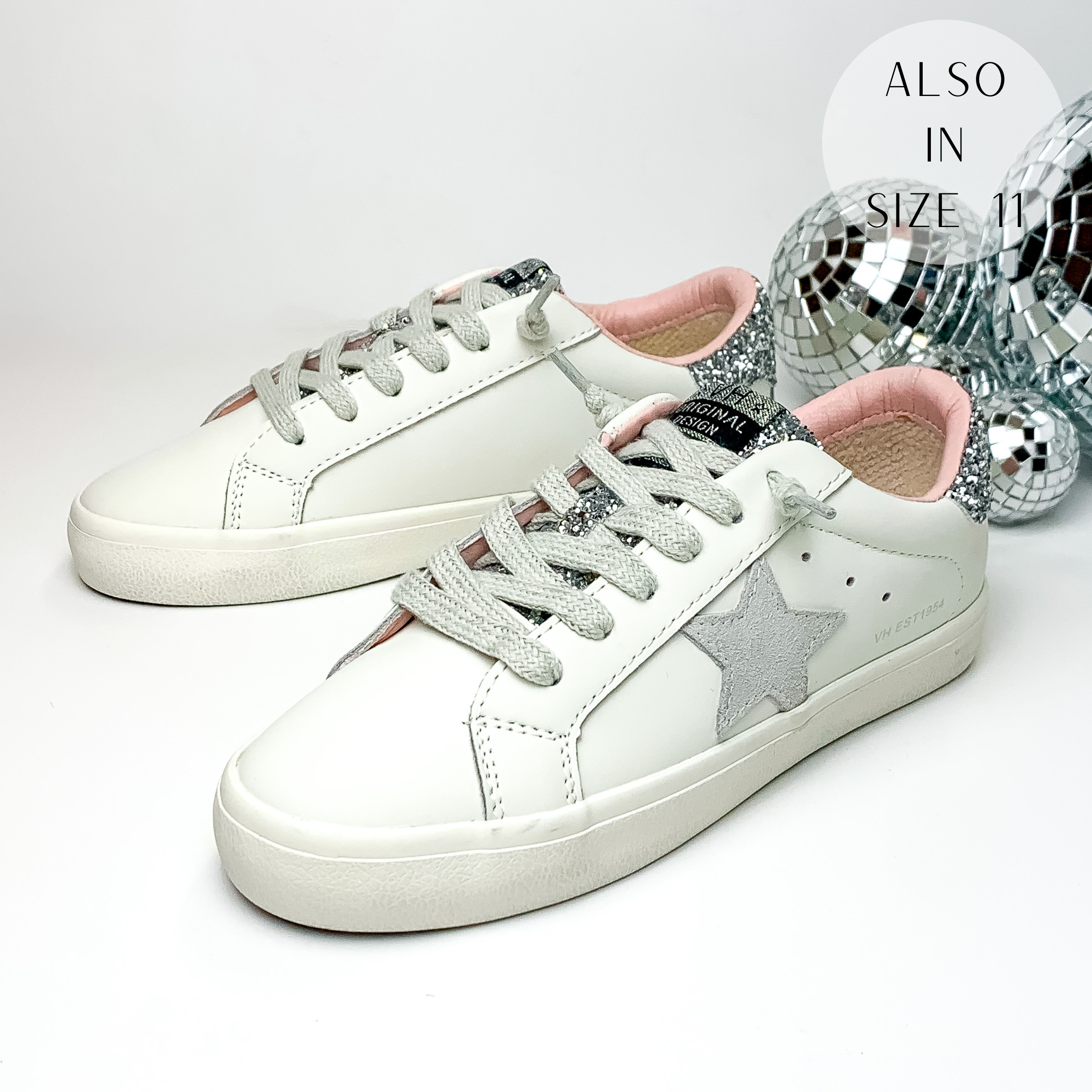 White tennis shoes with silver glitter on the tongue and on the back of the heel. These shoes also have a light grey star emblem on the side of the shoe. These shoes are pictured on a white background with disco balls behind them on the right hand side.
