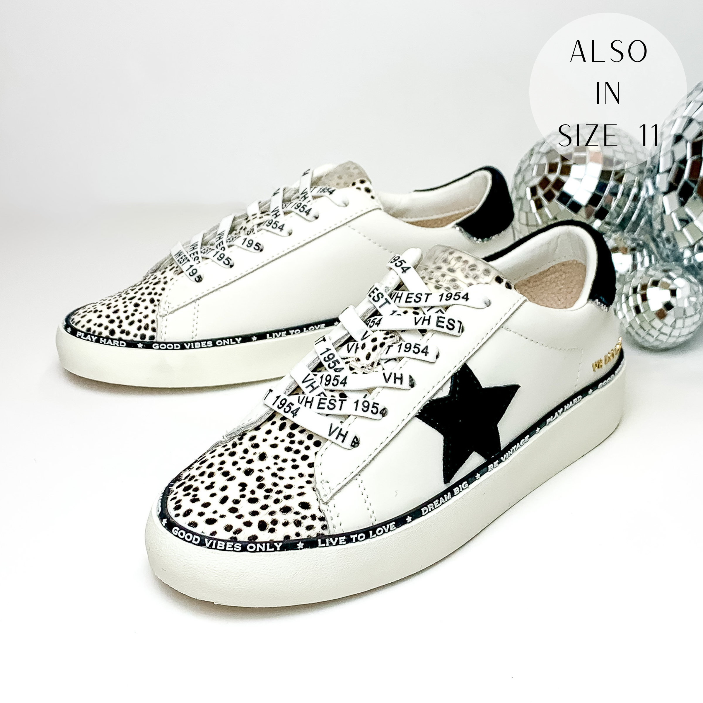 White tennis shoes with a black cheetah print on the tongue down to the toe and a black heel patch with a silver outline. These shoes also include a black rubber outline of the shoe that has positive phrases on it and they have a black star emblem on the side of the shoe. These shoes are pictured on a white background with disco balls behind them on the right hand side.