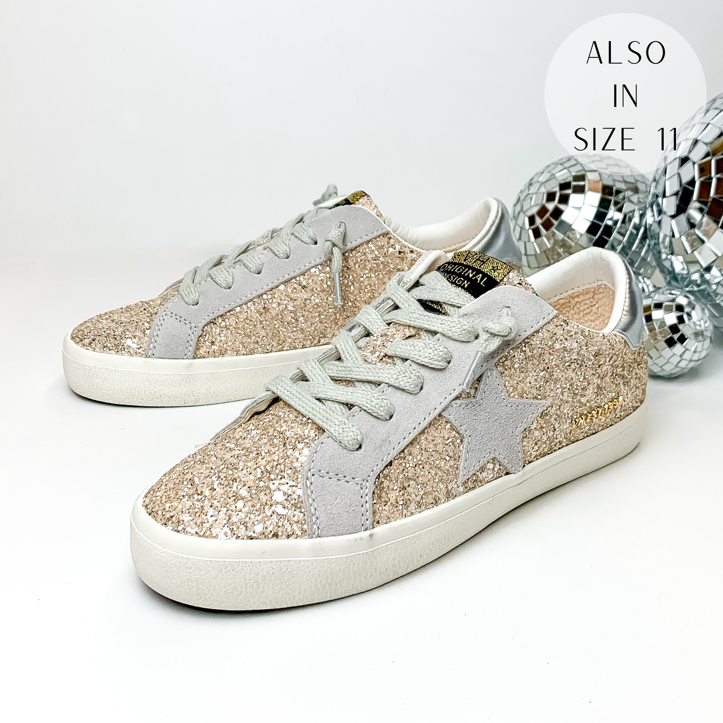 Pictured are tennis shoes with a champagne glitter inlay with light grey accents. These shoes also include a metallic silver patch on the heel and they have a light grey star emblem on the side of the shoe. These shoes are pictured on a white background with disco balls behind them on the right hand side.