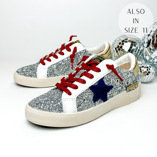Pictured are tennis shoes covered in a silver glitter. These shoes also include a metallic gold patch on the heel, a navy star emblem on the side of the shoe, and red shoe laces. These shoes are pictured on a white background with disco balls behind them on the right hand side.