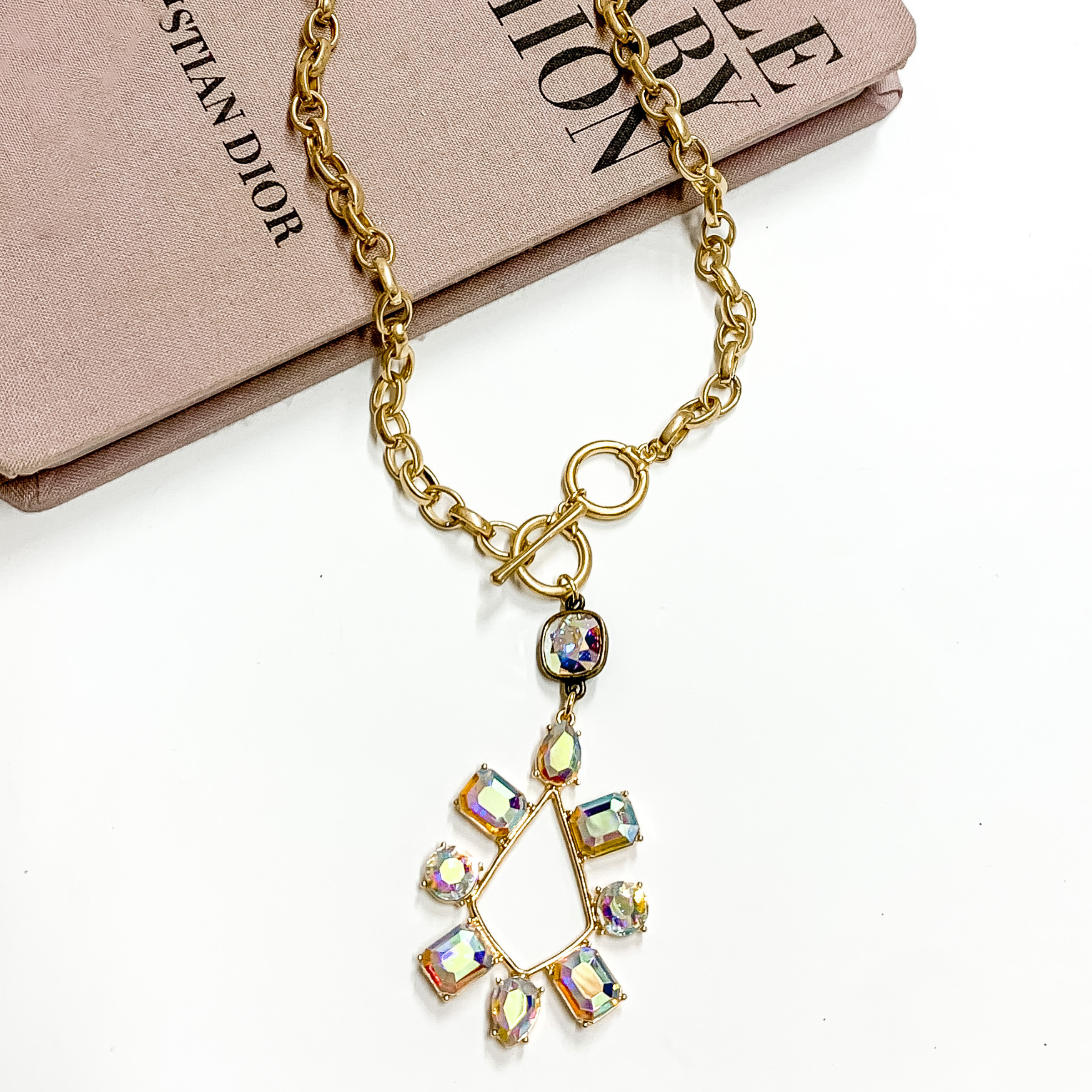 Gold chain necklace with toggle, front clasp and a multi shape ab crystal teardrop pendant. This necklace is pictured laying partially on a mauve colored book on white background. 