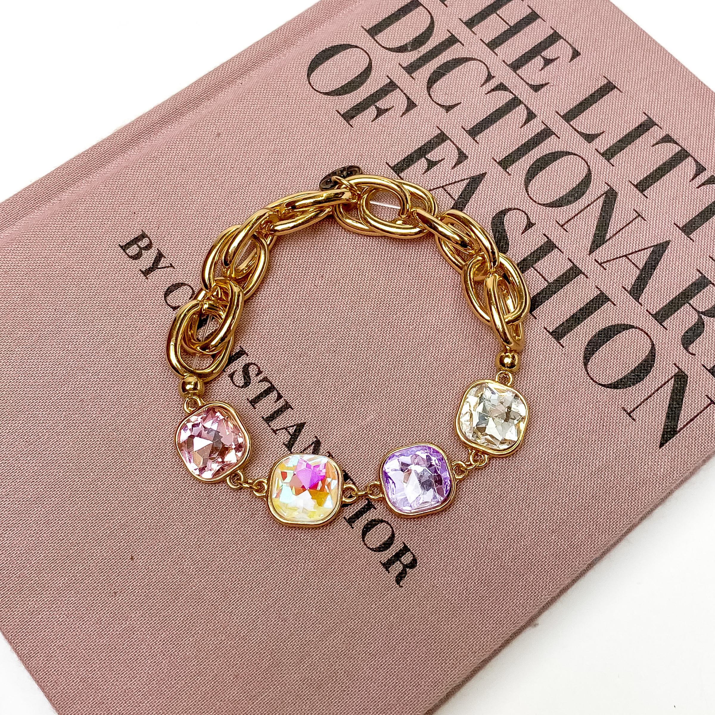 Gold Chain bracelet with four cushion cut crystals. These crystals come in light pink, opal ab, lavender, and clear. This bracelet is pictured on a mauve colored book on a white background. 