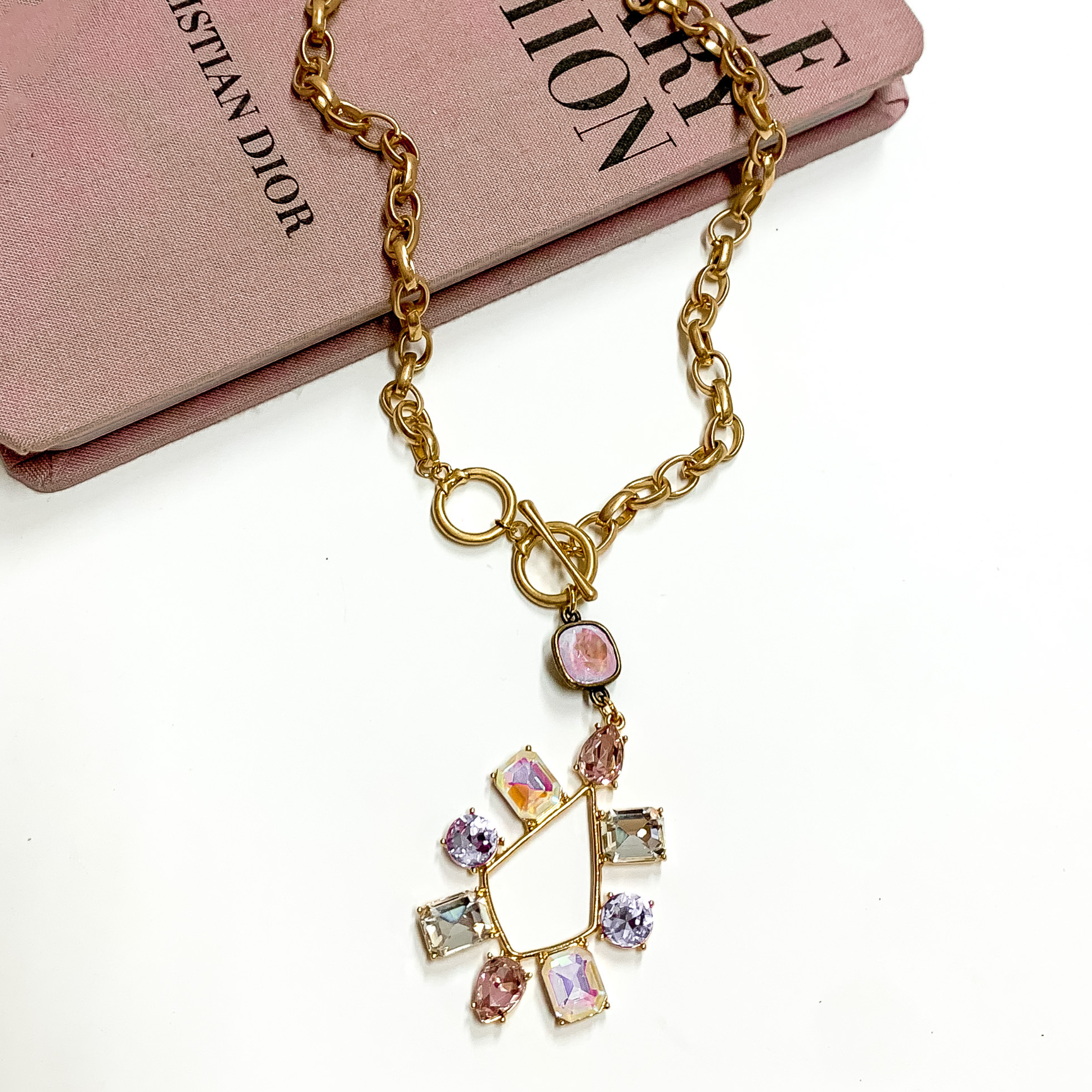 Gold chain necklace with toggle, front clasp with a lavender cushion cut crystal drop and a multi color crystal teardrop. This necklace is pictured laying partially on a mauve colored book on a white background. 