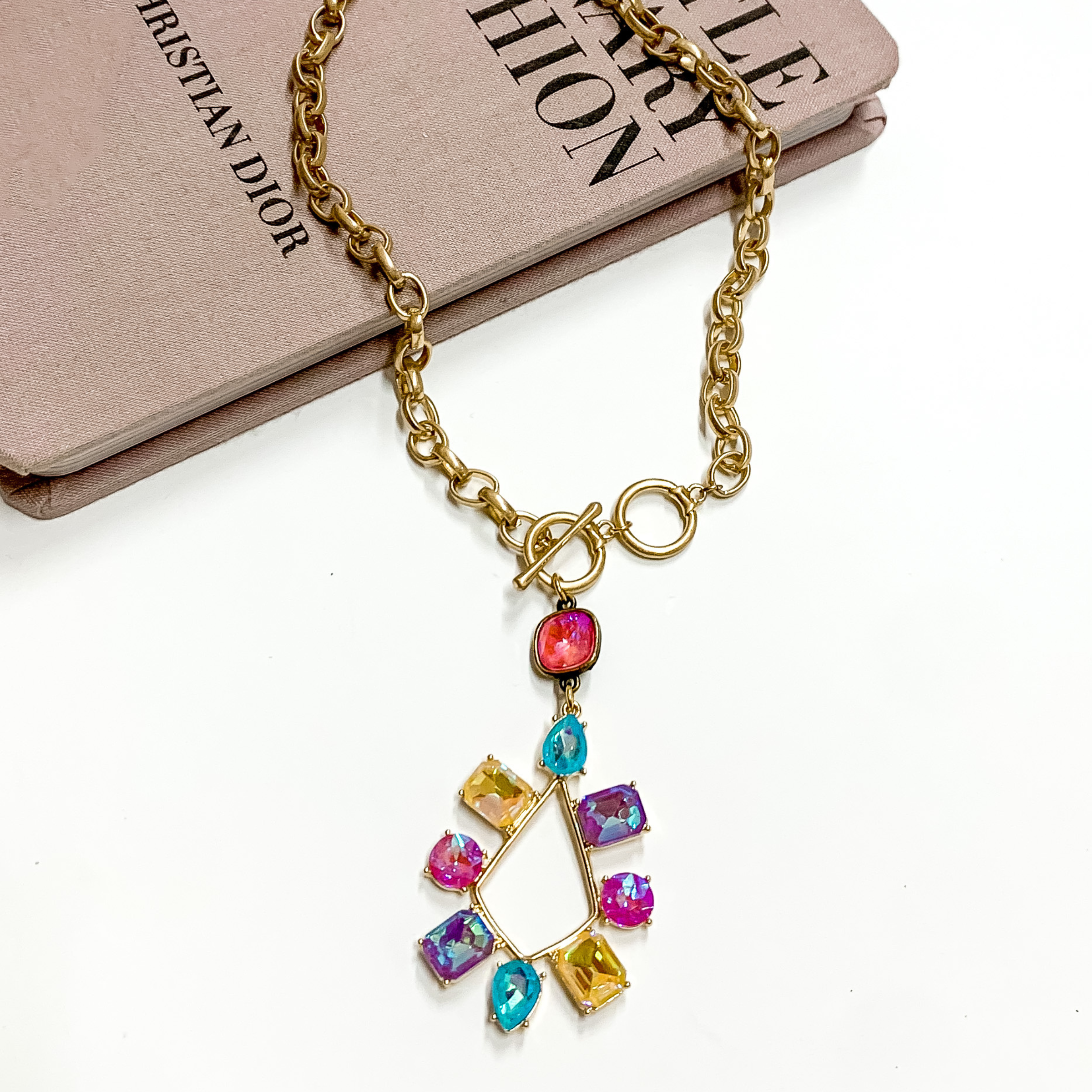 Gold chain necklace with toggle, front clasp with a pink cushion cut crystal drop and a multi color crystal teardrop. This necklace is pictured laying partially on a mauve colored book on a white background. 
