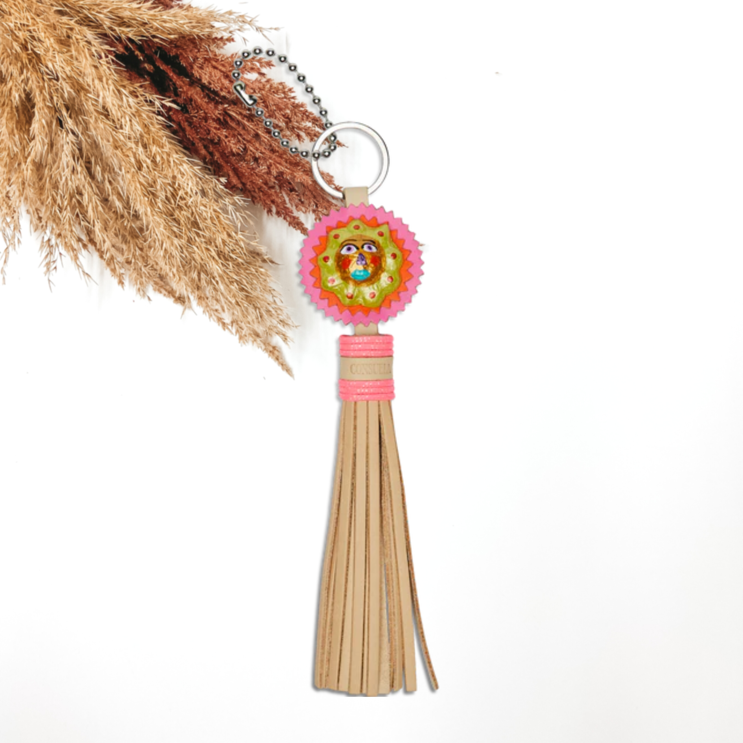 Silver key ring with a pink, circle charm and light tan tassels. The circle charm has a multicolored face in the center. This charm is pictured on a white background with pompous grass in the top right corner.  