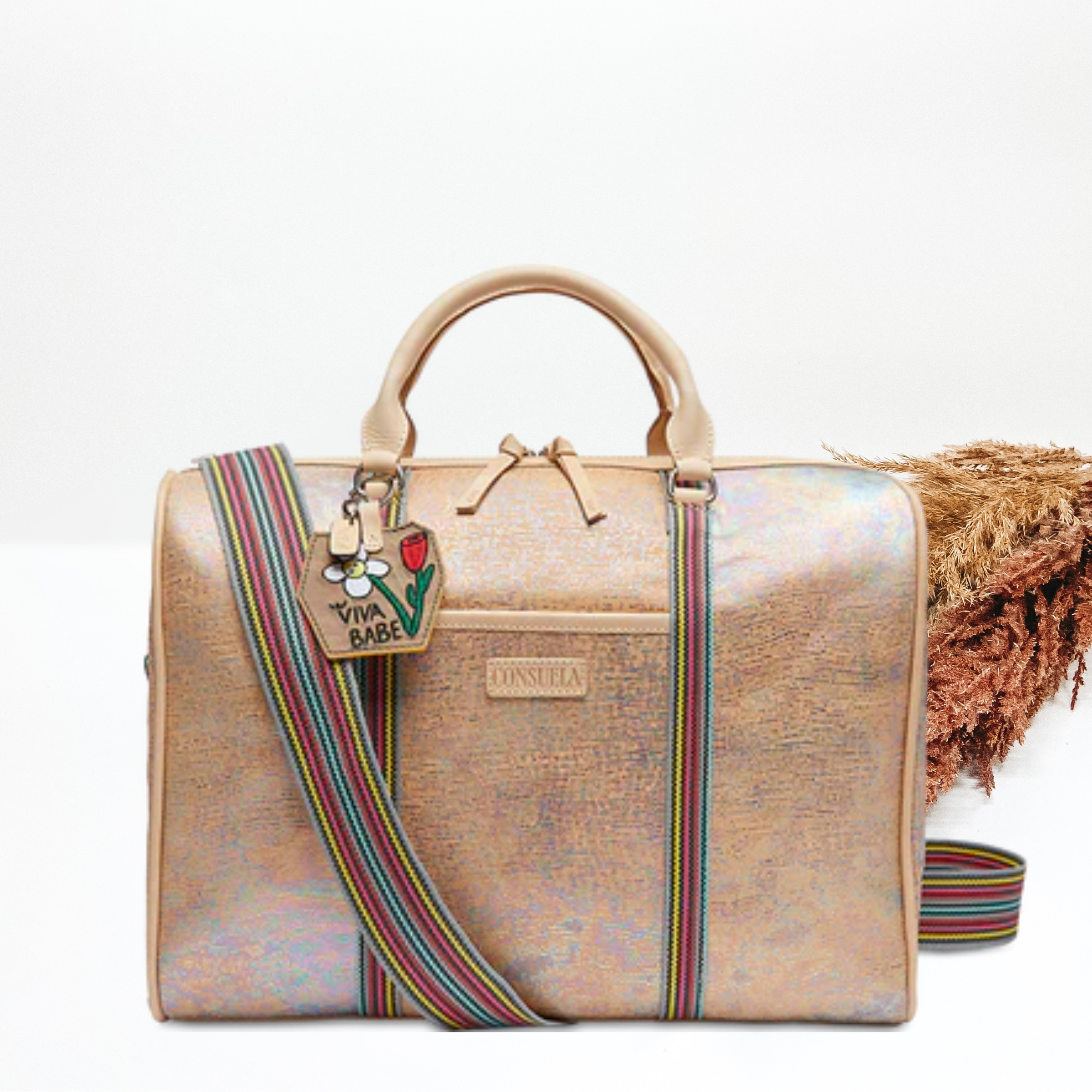 Pictured is a duffle bag with iridescent, multicolor design. This duffle bag includes short, light tan handles and a long, striped strap. This bag is pictured on a white background with pompous grass on the left side of the picture.