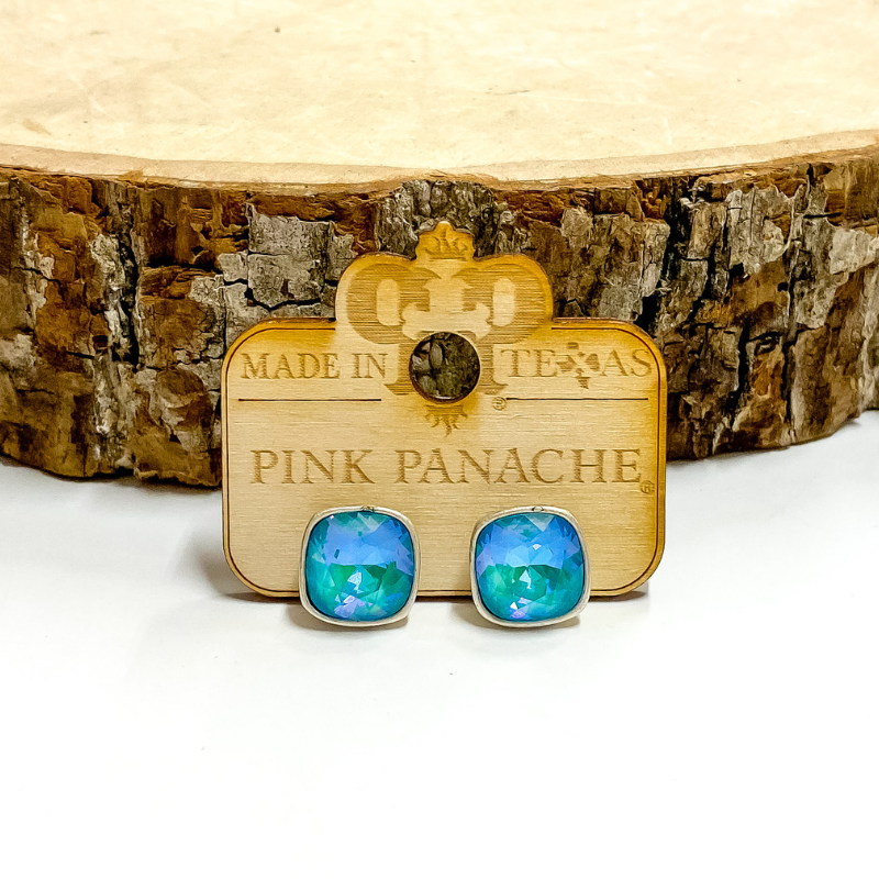 A pair of silver, square stud earrings with laguna delight cushion cut crystals. These earring are pictured on a wood holder on a white background with a piece of wood in the background.