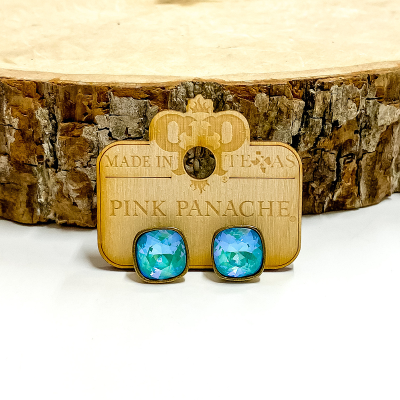 A pair of bronzr, square stud earrings with laguna delight cushion cut crystals. These earring are pictured on a wood holder on a white background with a piece of wood in the background.