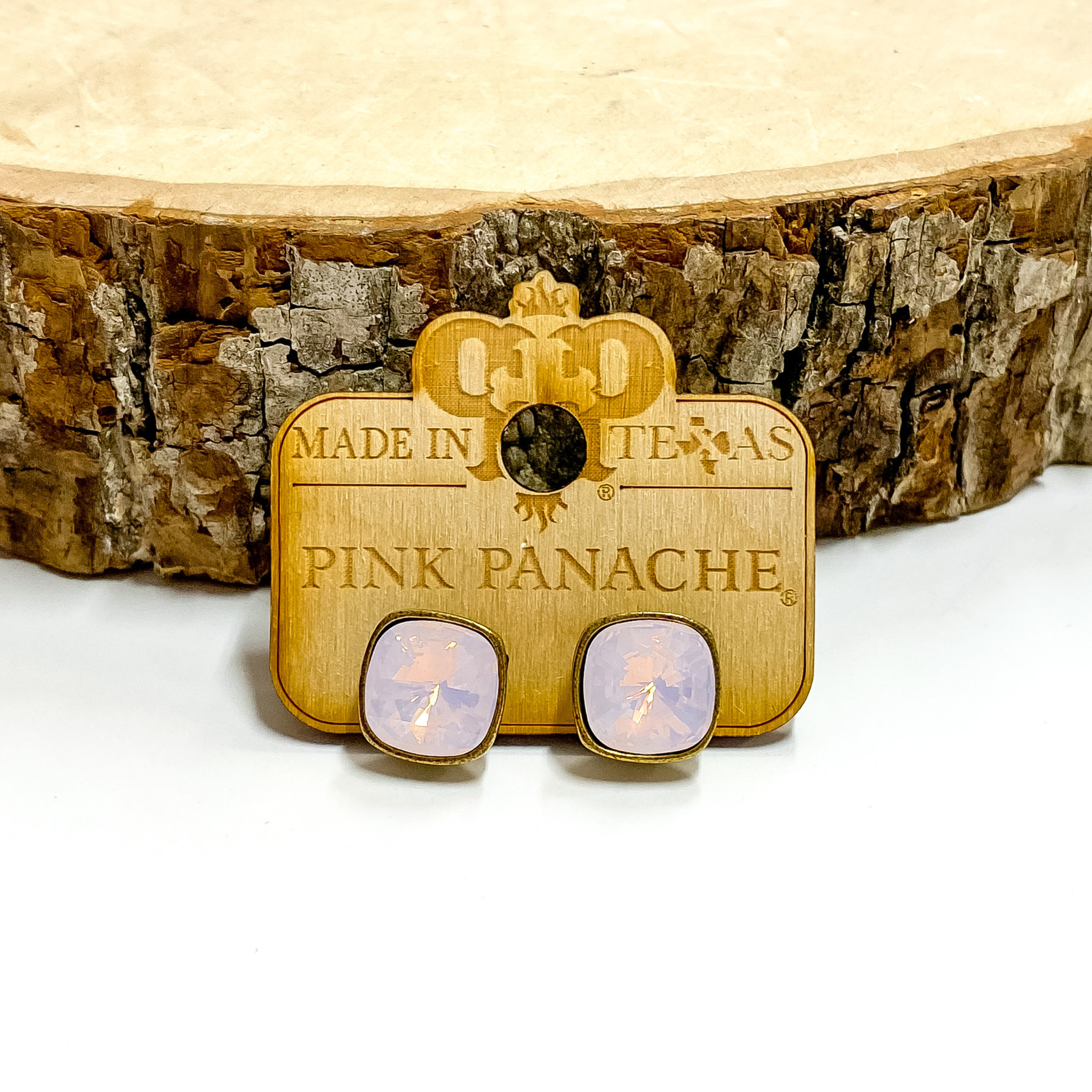 A pair of bronze, square stud earrings with rose water opal cushion cut crystals. These earring are pictured on a wood holder on a white background with a piece of wood in the background.