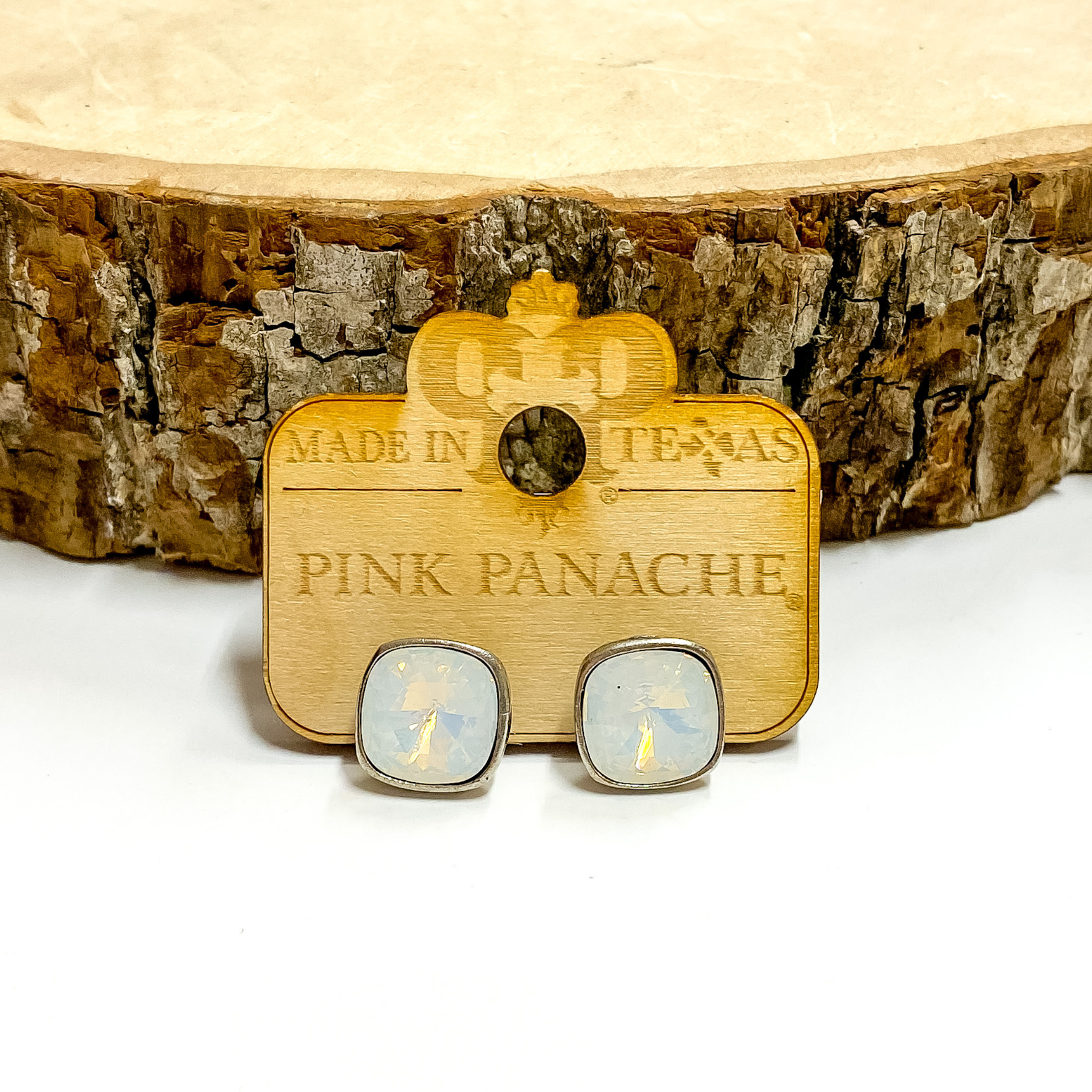 A pair of silver, square stud earrings with water opal cushion cut crystals. These earring are pictured on a wood holder on a white background with a piece of wood in the background.