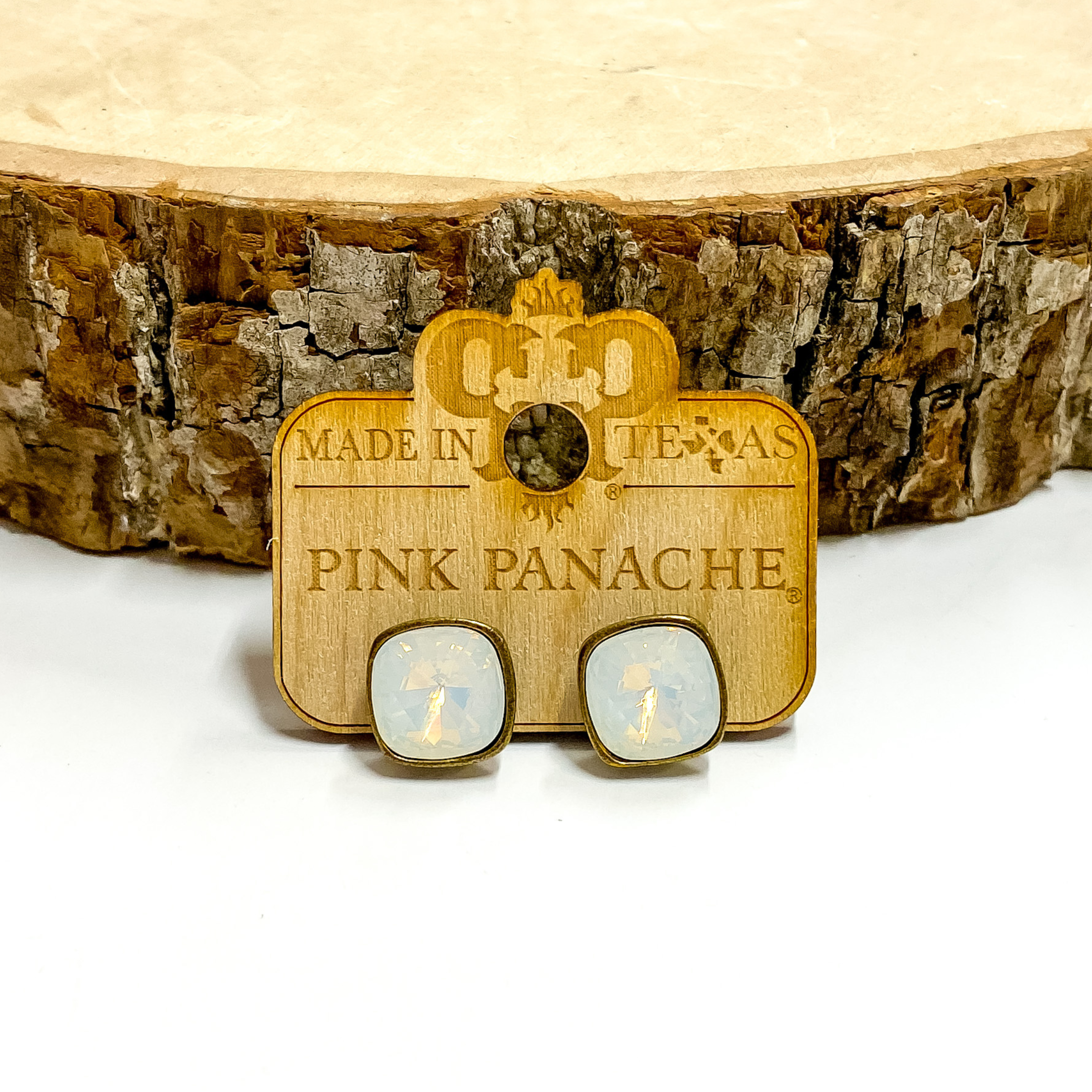 A pair of bronze, square stud earrings with water opal cushion cut crystals. These earring are pictured on a wood holder on a white background with a piece of wood in the background.
