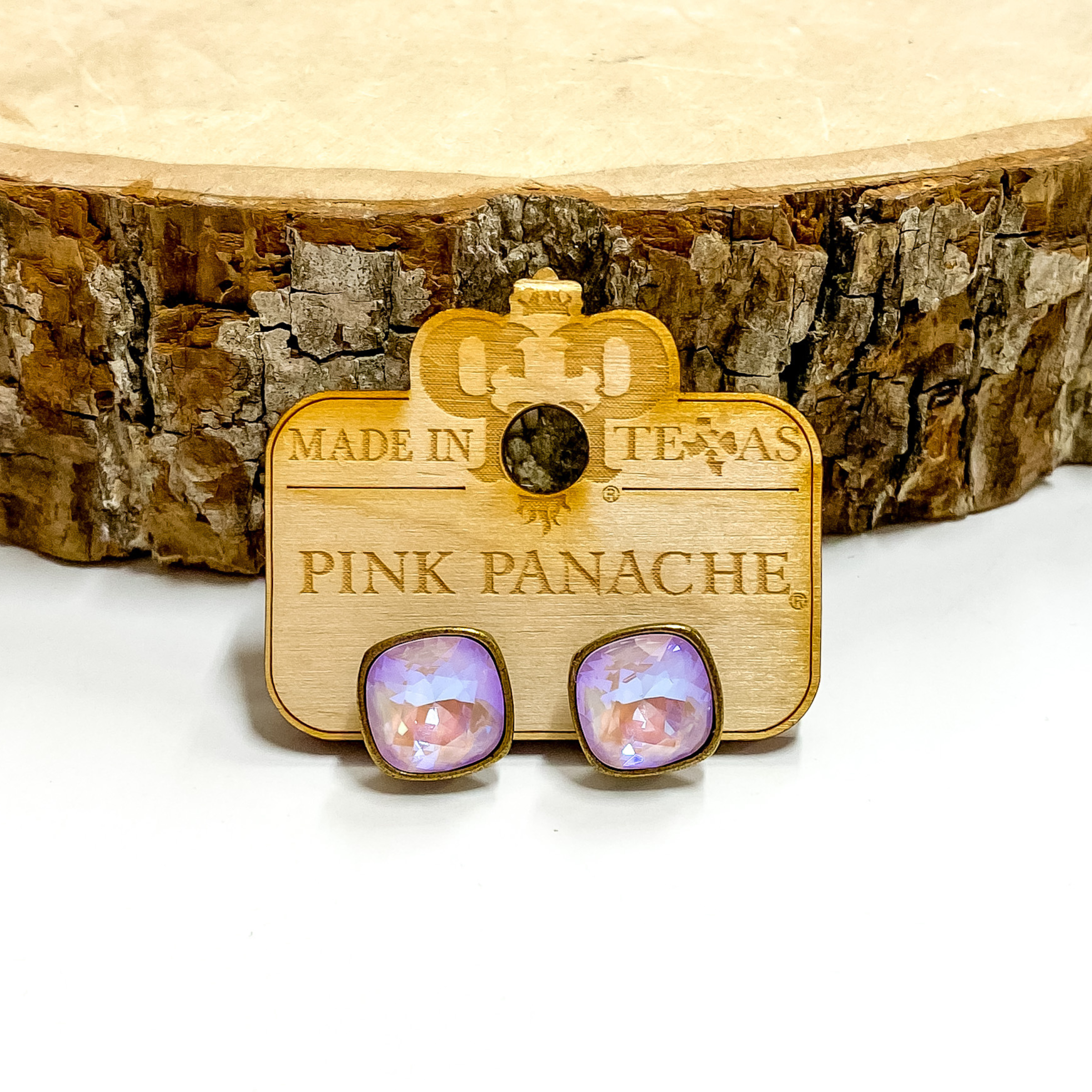A pair of bronze, square stud earrings with lavender cushion cut crystals. These earring are pictured on a wood holder on a white background with a piece of wood in the background.