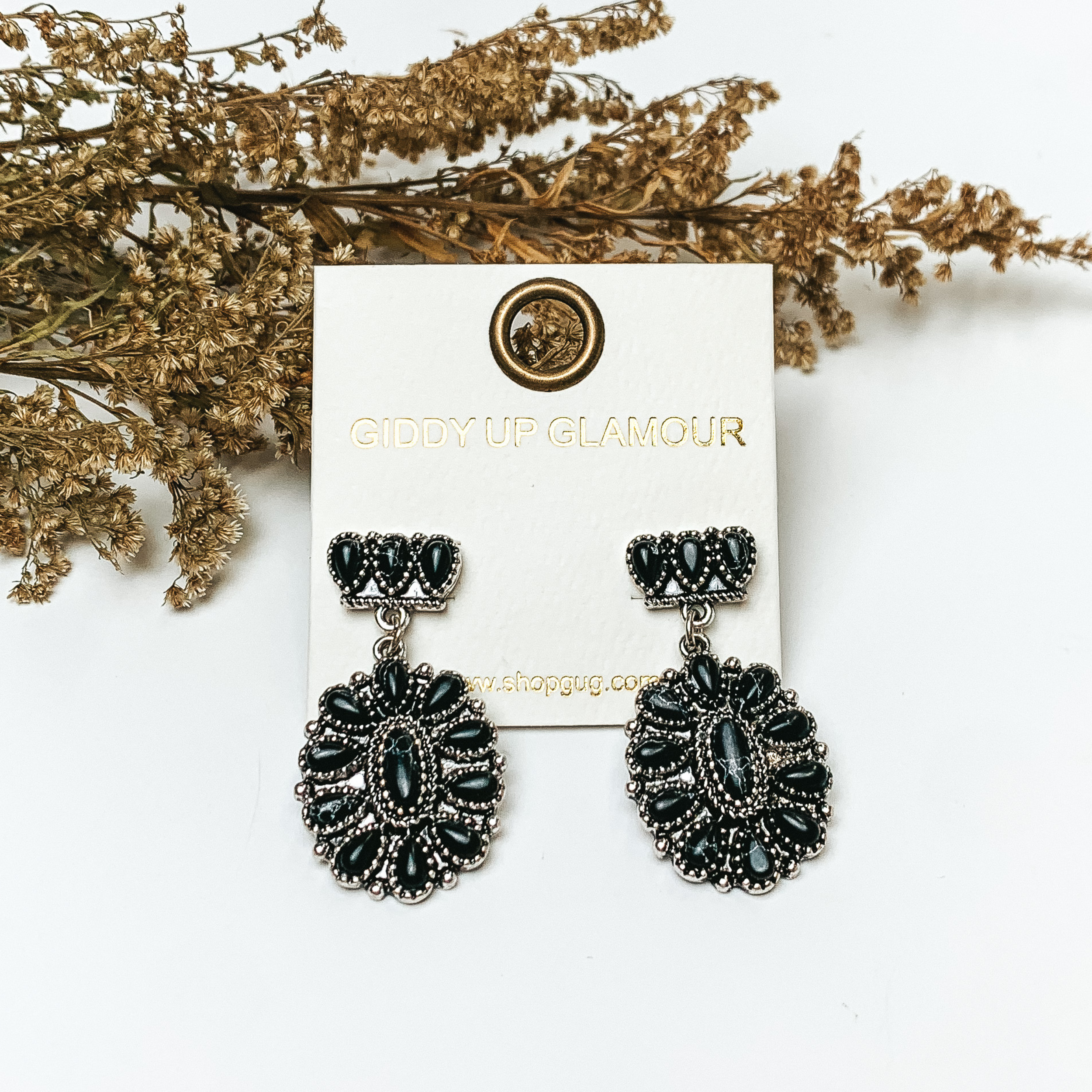 Silver tone cluster post earrings with oval cluster drop with black stones. These earrings are pictured on a white background with greenery behind the earrings.